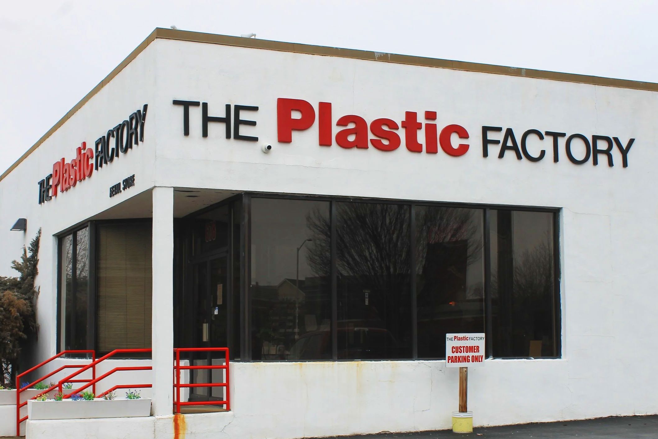 Exterior of The Plastic Factory retail store with a large sign on the white building, featuring the words 'The Plastic Factory' with 'Plastic' in red and the rest in black. A small sign in front reads 'Customer Parking Only'.