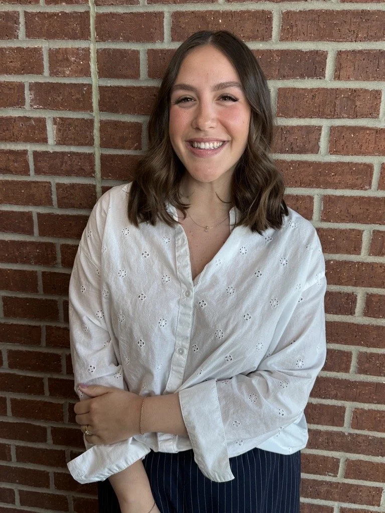 A young woman with shoulder-length brown hair smiling while standing in front of a brick wall, wearing a white eyelet blouse and dark striped pants.