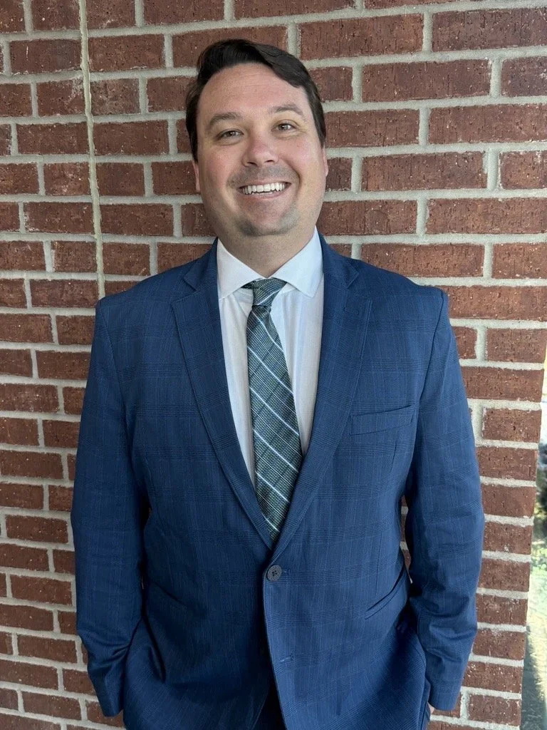 Man in a blue suit and striped tie standing in front of a brick wall, smiling.