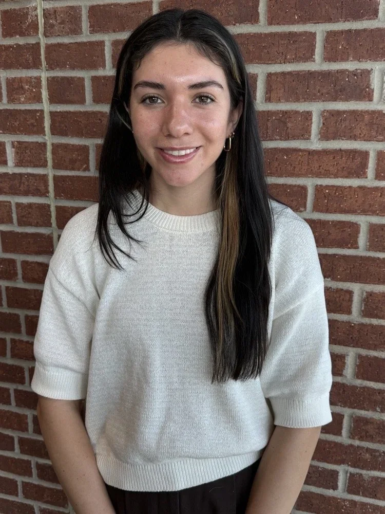 A young woman with long dark hair and light skin, smiling, standing against a red brick wall. She is wearing a white sweater and black pants.