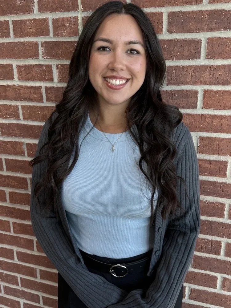 A young woman with long, wavy dark hair smiling and standing in front of a brick wall, wearing a light blue top, a dark gray cardigan, and a necklace.