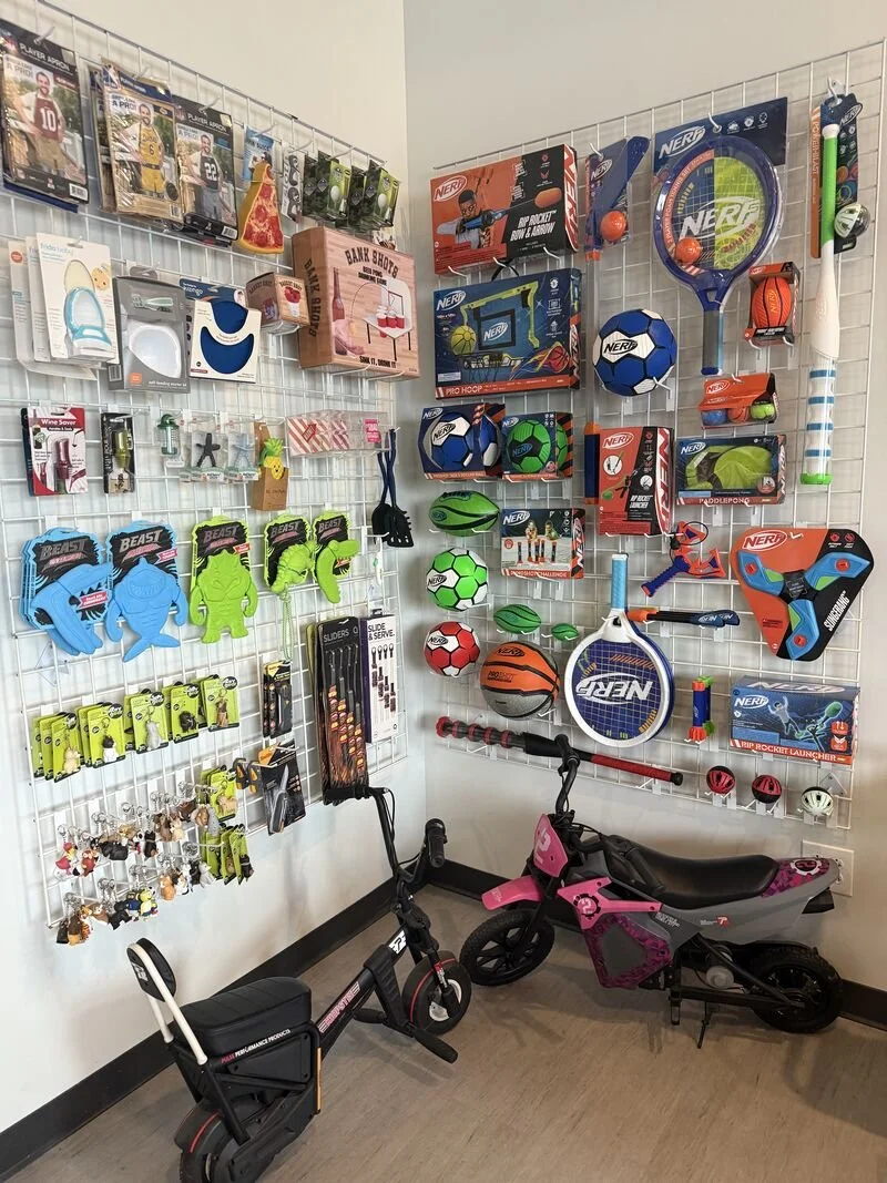 A corner display of various children's toys and accessories, including sports balls, a tennis racket, a small bicycle, a scooter, and a toy motorcycle, with shelves and hooks holding toy packaging and accessories.