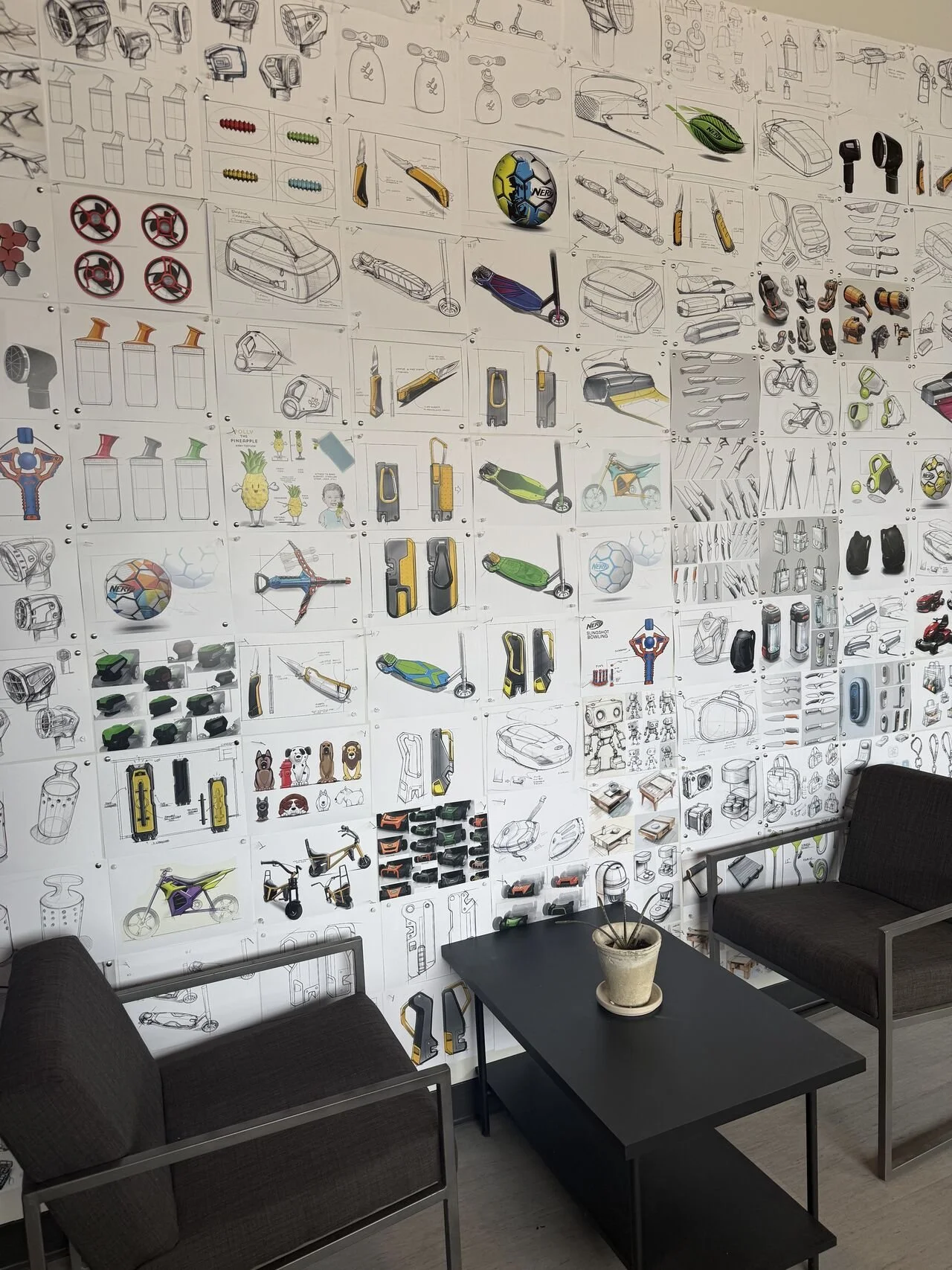 Wall filled with technical sketches and concept art of various sports equipment, tools, and gadgets, including skateboards, bikes, scooters, and accessories, in a modern workspace with black chairs and a table with a plant.