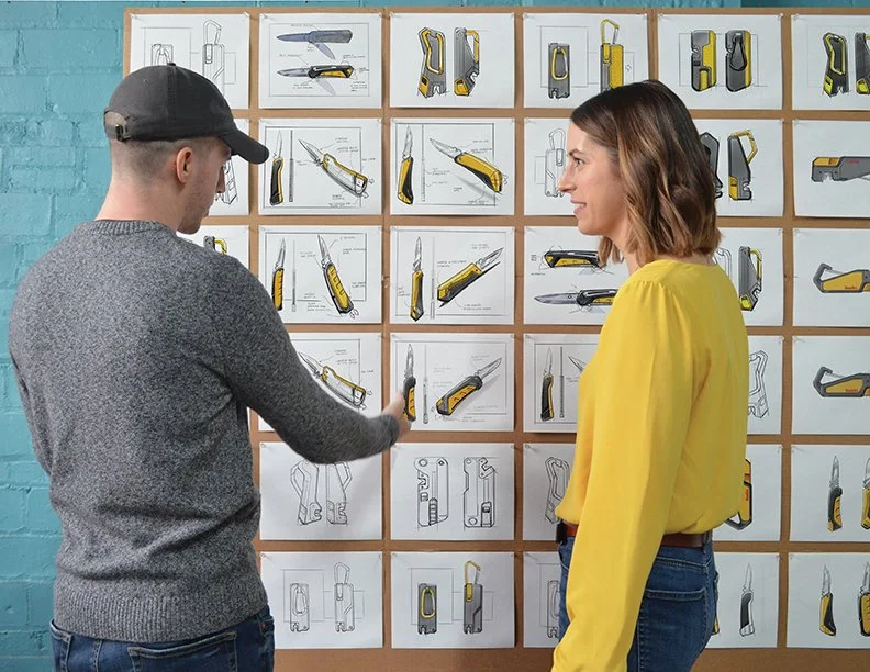 Two people, a man and a woman, looking at a display of industrial design sketches on a wall