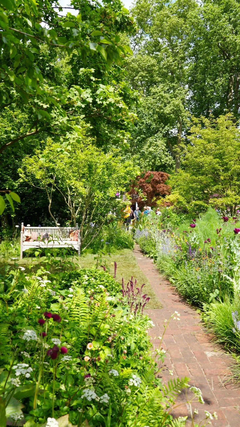 A brick path leading to a white bench between full perennial borders under trees