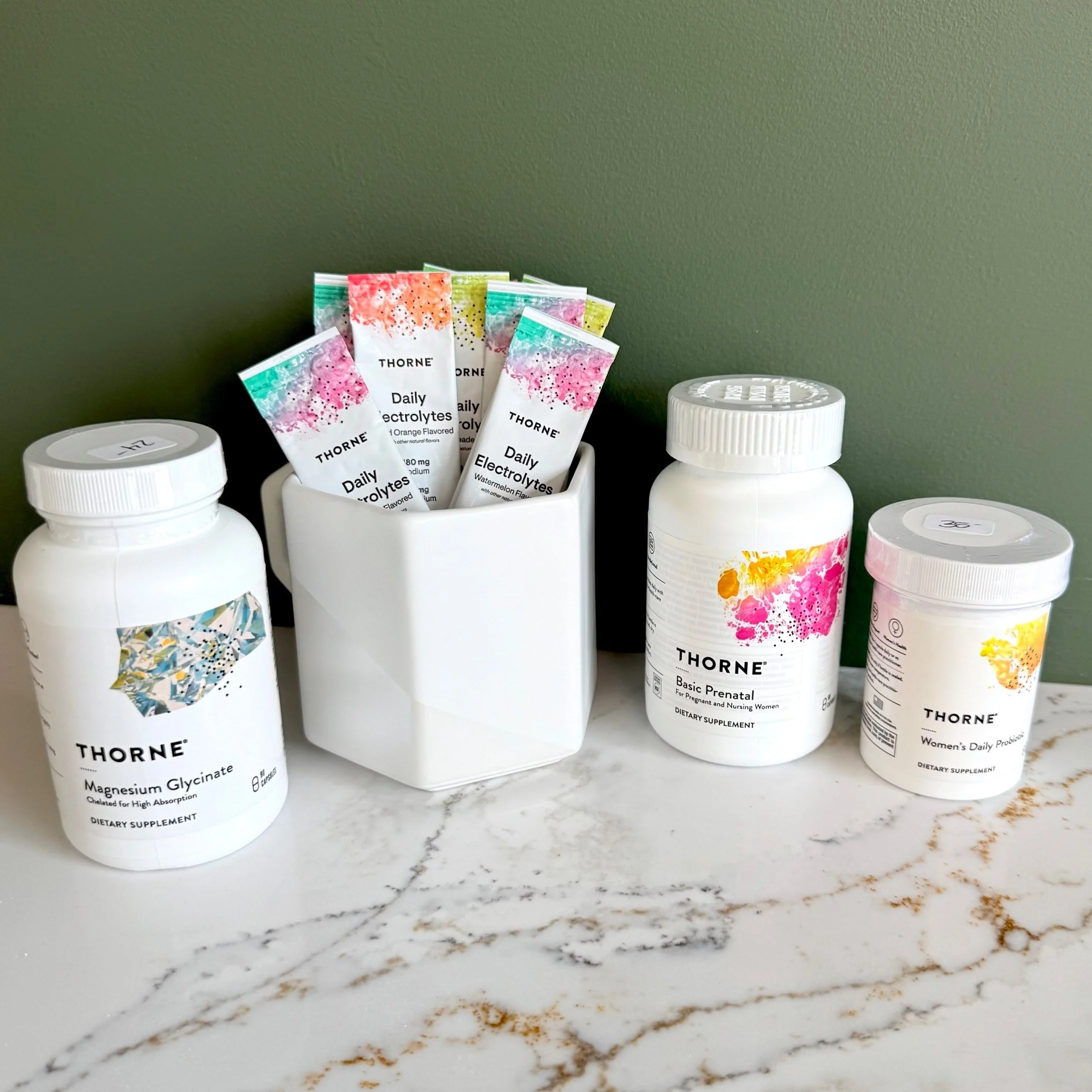 Assorted dietary supplement bottles and packets on a marble countertop against a green wall, including magnesium glycinate, prenatal vitamins, women's daily probiotics, and electrolyte packets.