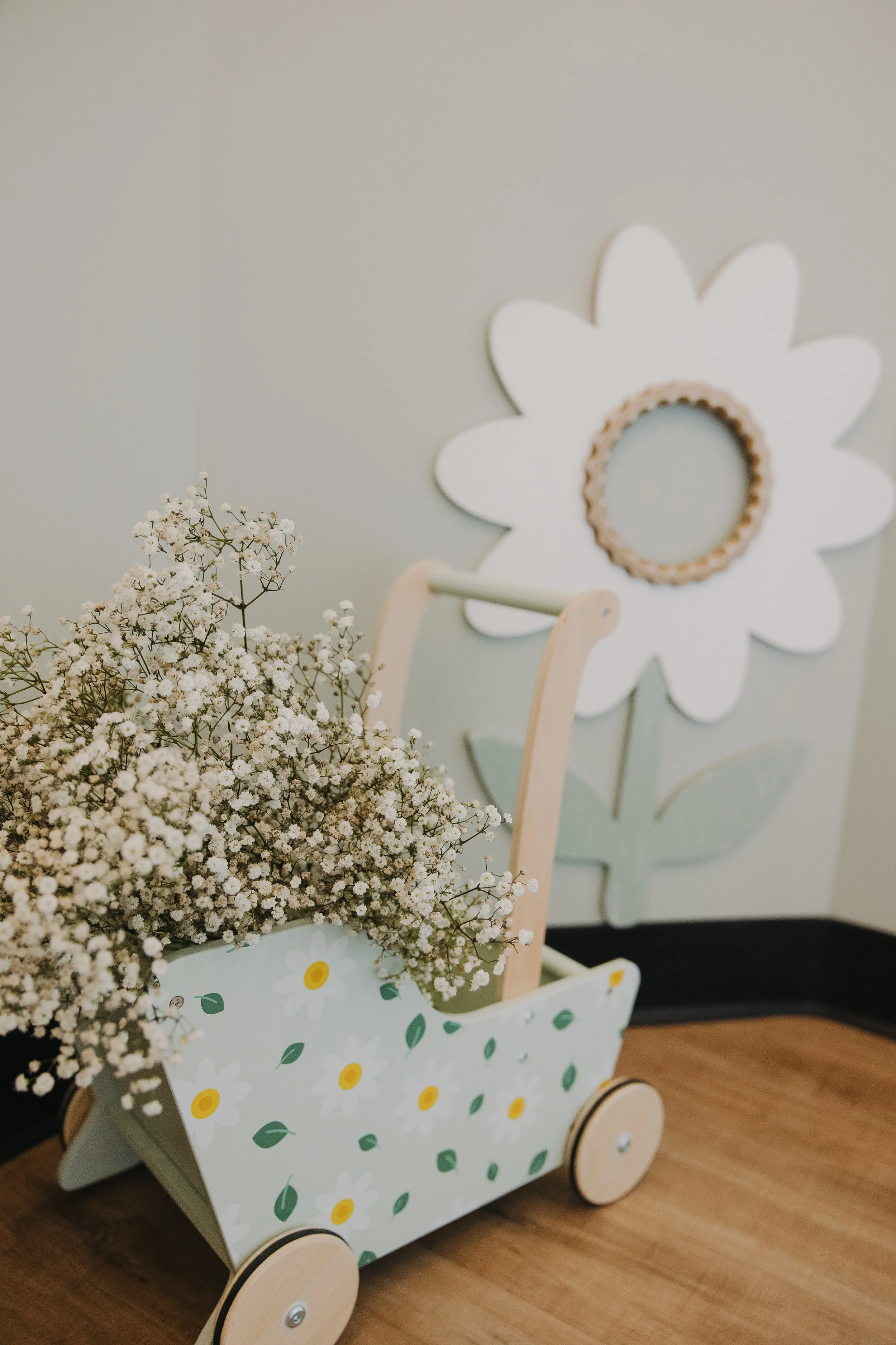 A decorative wooden flower-shaped mirror on the wall, a wooden toy wagon with a floral pattern on it, filled with white baby's breath flowers, placed on a hardwood floor.