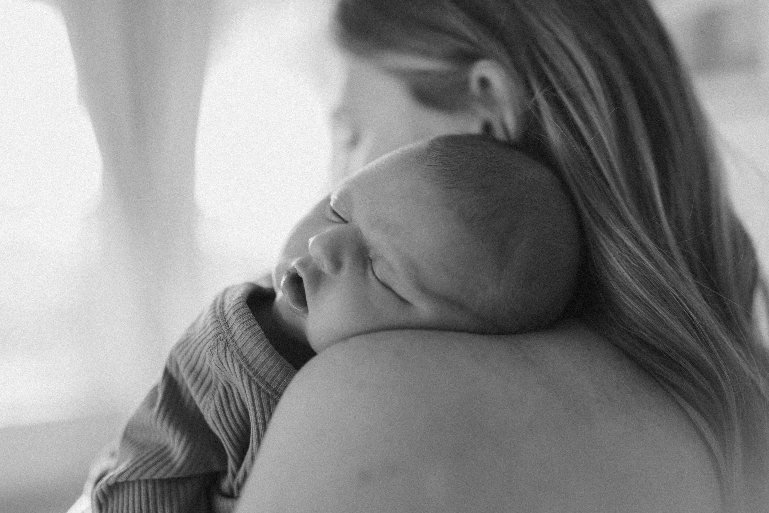 Black and white photograph of a sleeping newborn baby resting on a woman's shoulder, with the woman gently holding the baby.