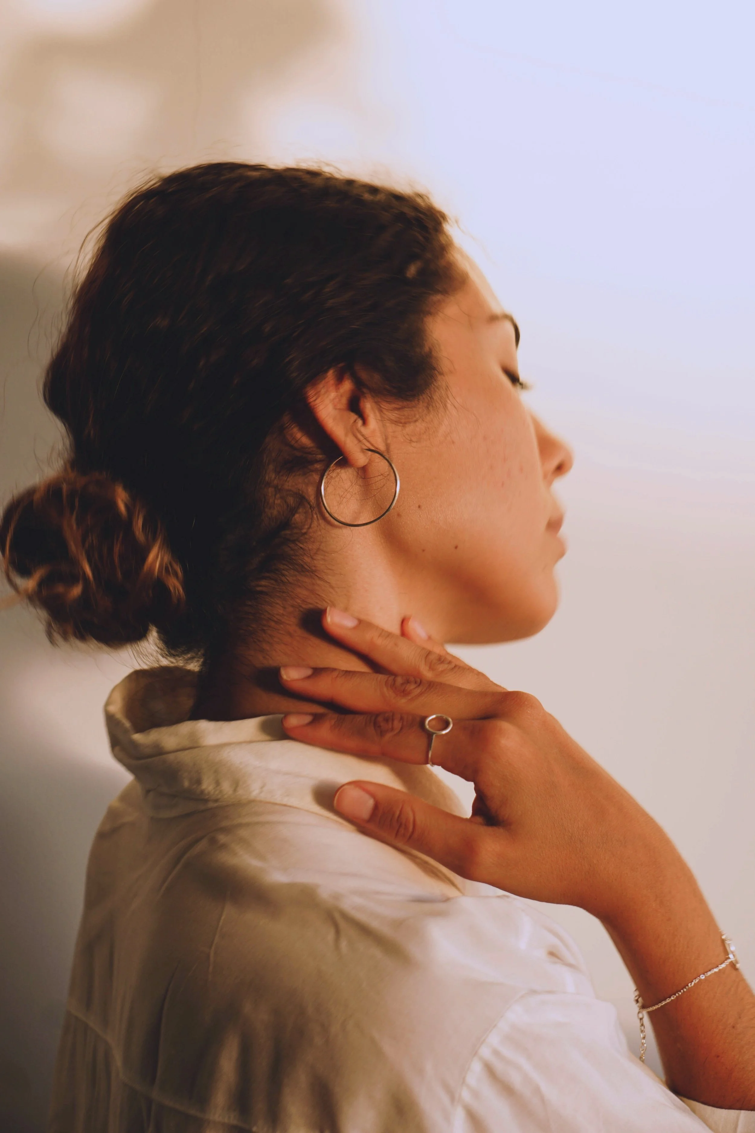 Side profile of a woman with a bun hairstyle, wearing hoop earrings, a ring, and a bracelet, resting her hand on her neck with her eyes closed.