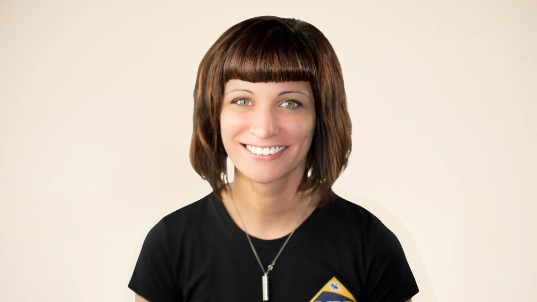 Woman with shoulder-length brown hair and bangs, wearing a black shirt with a graphic design and a necklace, smiling at the camera.