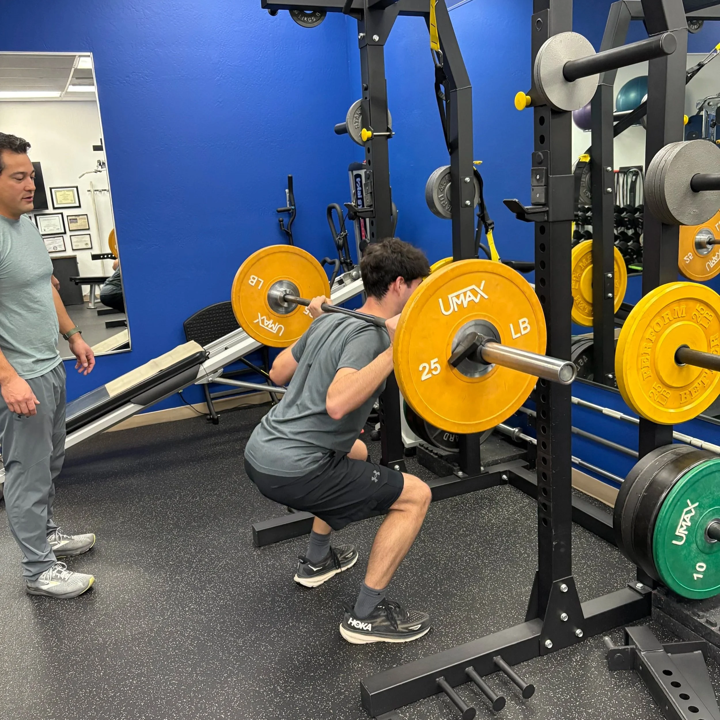 Teen athlete performs a barbell squat with a personal trainer supervising and coaching in a studio gym.