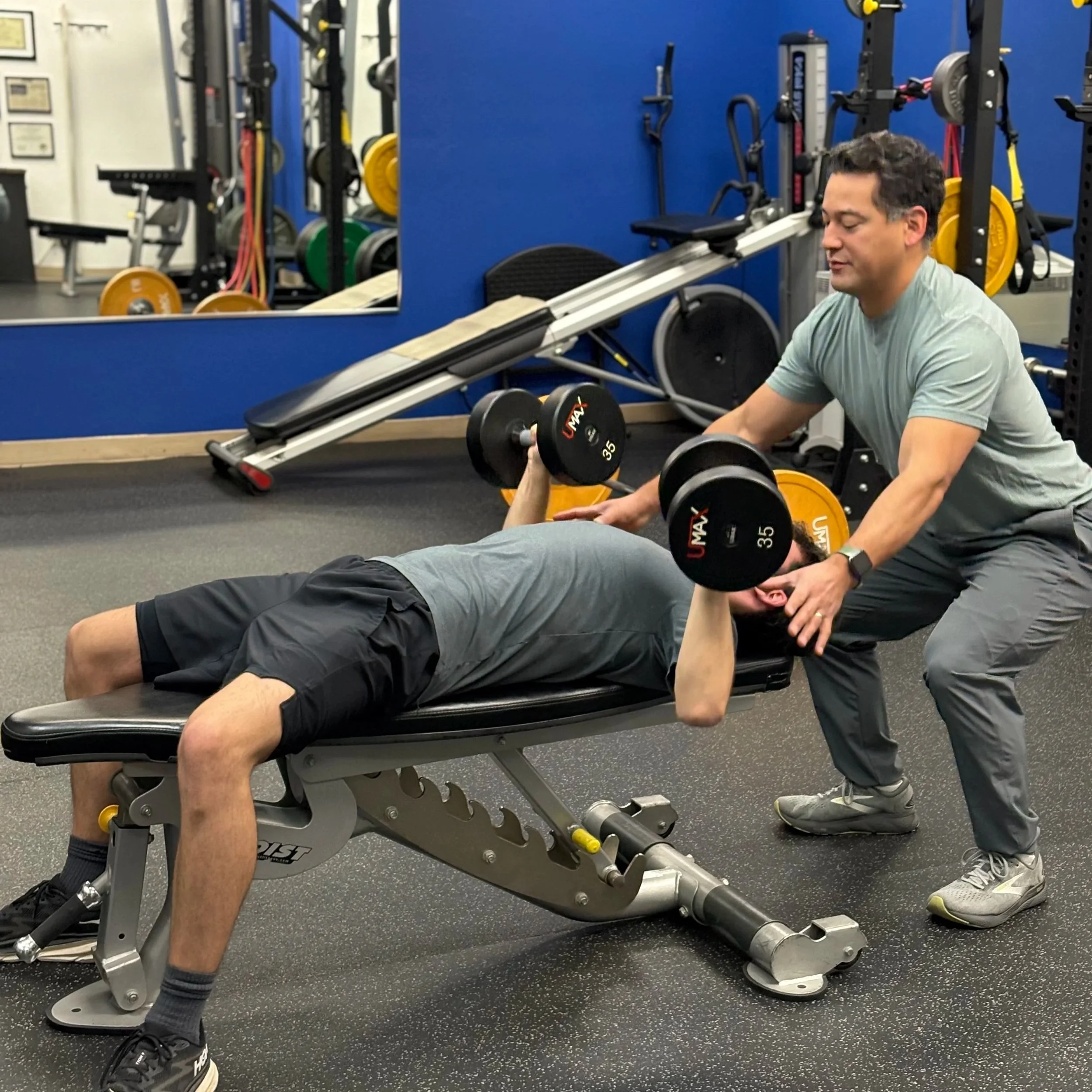 Teen athlete performing a dumbbell press while a personal trainer spots him in a gym studio.