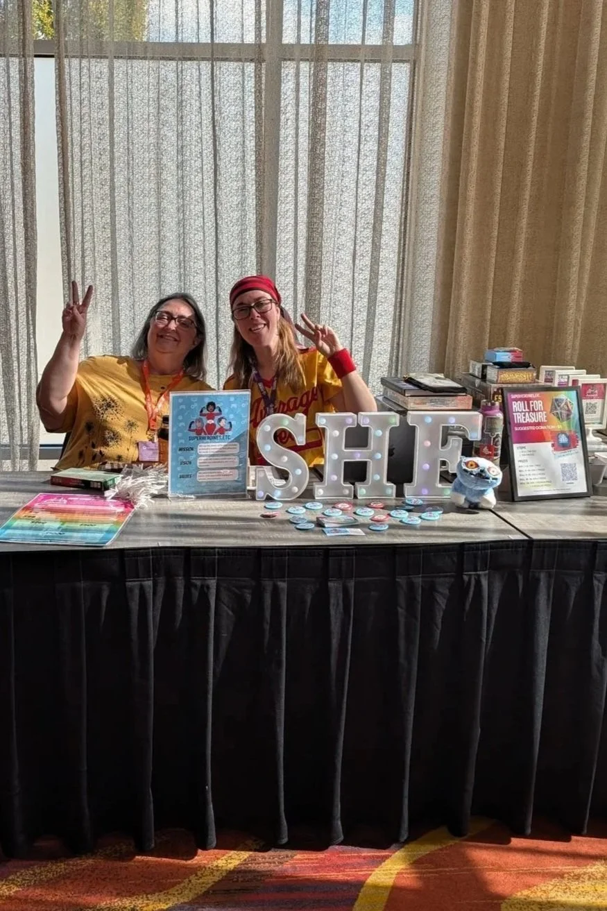 Two women sitting behind a table at a booth, smiling and flashing peace signs. The table displays promotional materials, a sign with the word 'SHIFT,' and various items, including round badges. The background features curtains and a window.