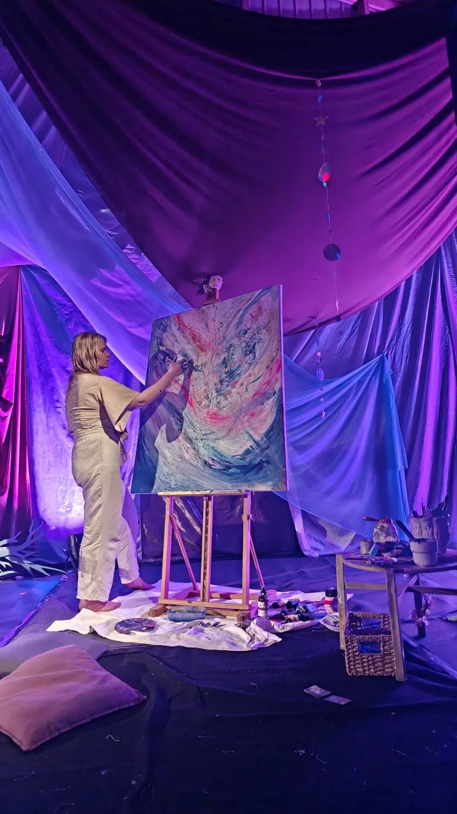 Nainen maalaa abstraktista taidetta verhoilun ja valaistuksen luomassa taidepajassa, jossa on seinät ja katto vaalean violetin ja sinisen sävyisinä. Nainen maalaa lavalla live maalausta tapahtumassa.