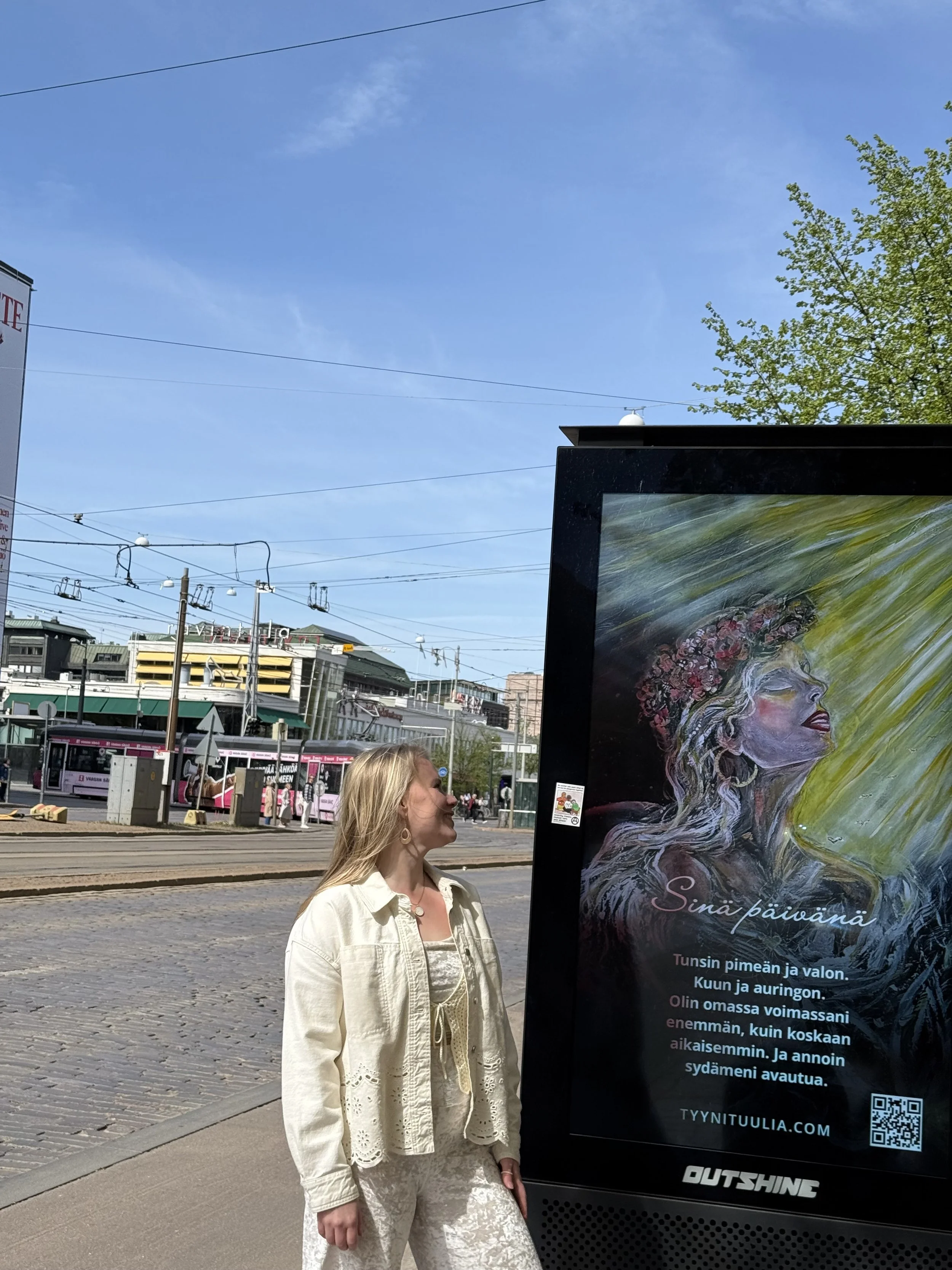Taiteilija Tuulia Kauppinen katsoo digitaalista mainonta taulua oikealla puolella ulkona kaupunkimaisemassa. Digimainonta taulussa on Kaikkeuden Jumalatar teos ja runo, oka alkaa sanoilla 'Sinä päivänä'.