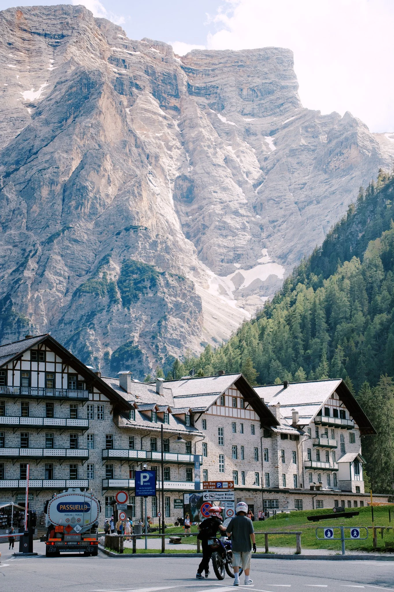 Scenic view of large stone building with alpine architecture, foregrounded by people and vehicles, set against a backdrop of towering, rugged mountains and dense green forest.