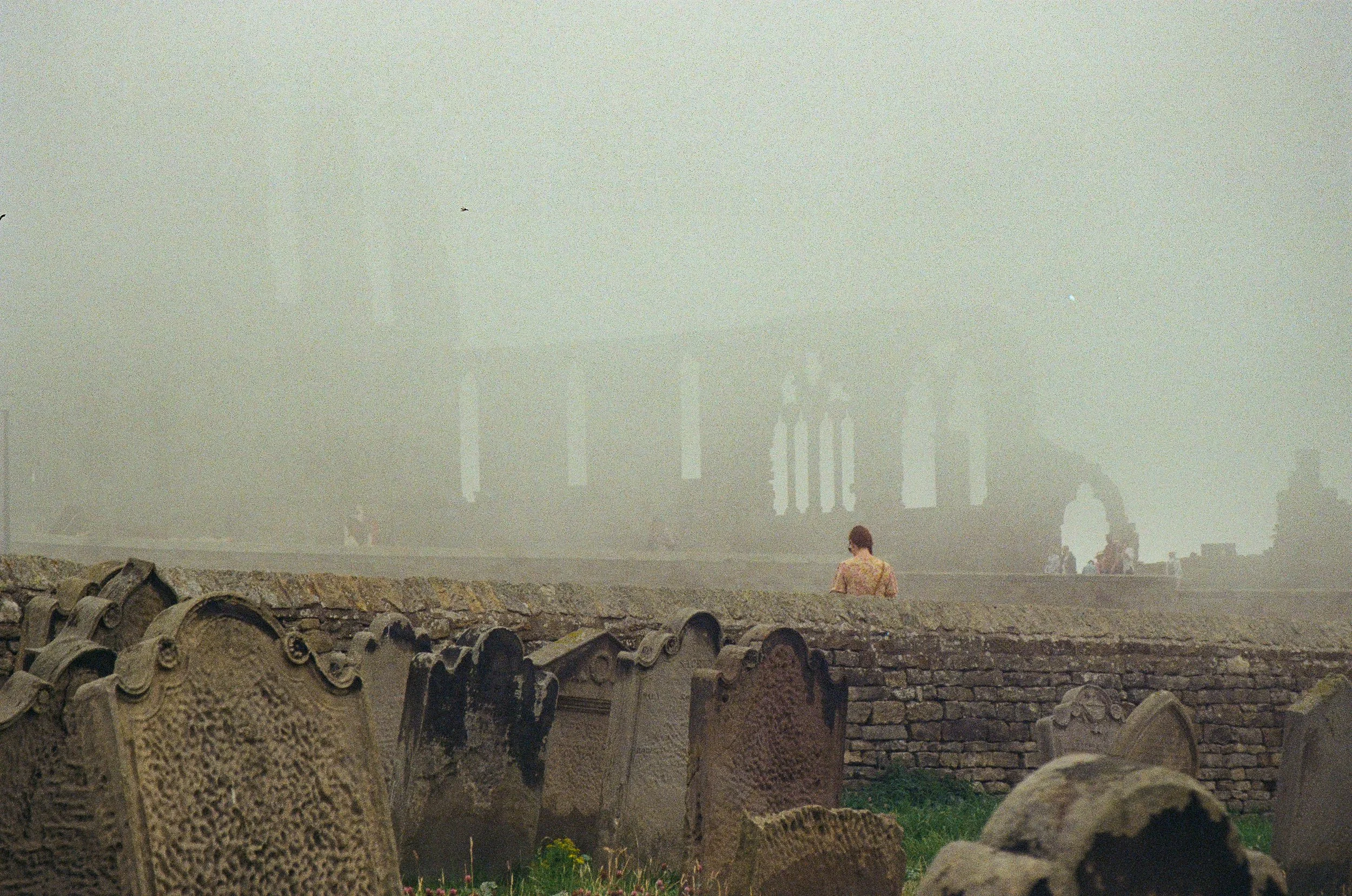 Foggy scene with old gravestones and ruins of a stone building in the background.