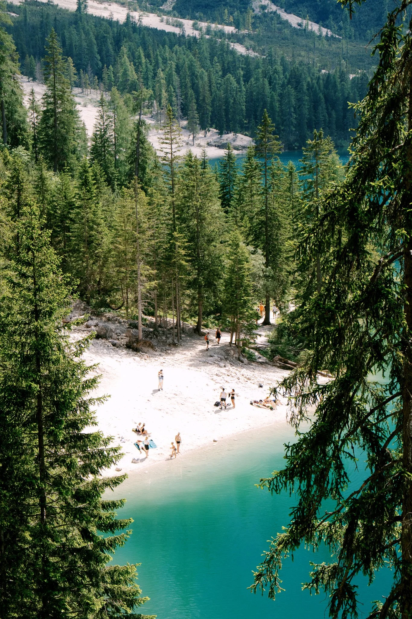 Scenic view of a turquoise lake surrounded by dense forest, with people relaxing on a sandy shore in a natural setting.