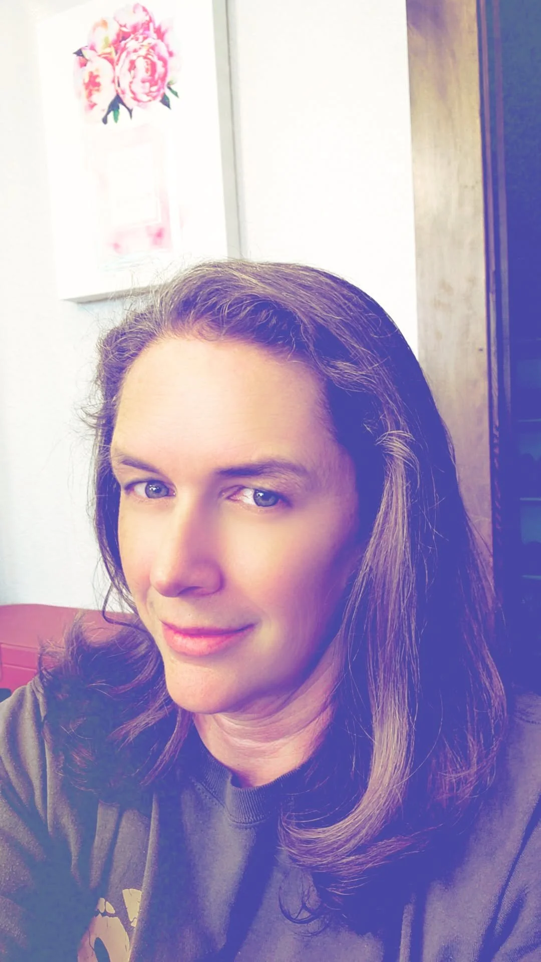 A woman with long brown hair and blue eyes taking a selfie indoors, with a floral painting on the white wall behind her.