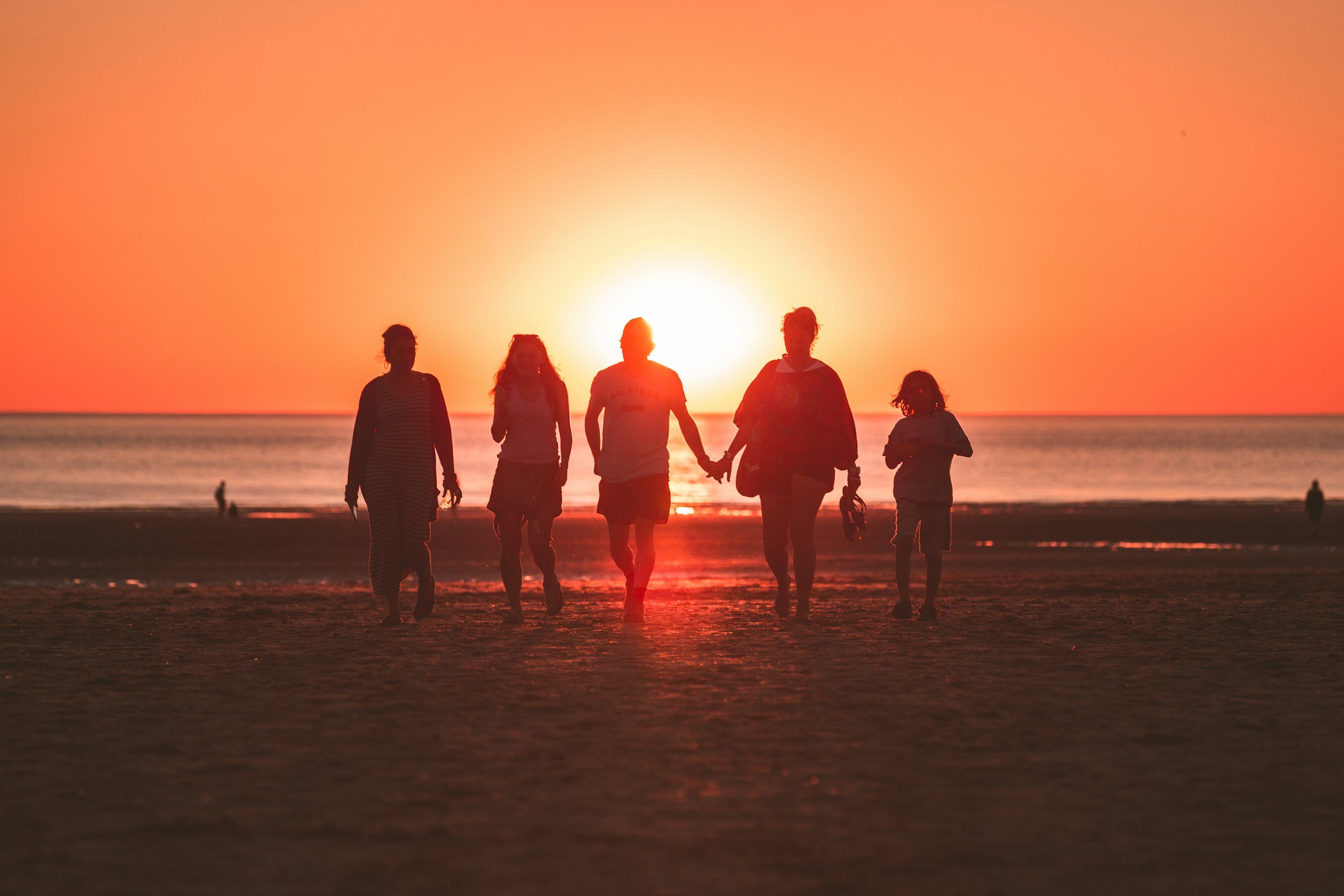 A group of five people walking hand in hand along the beach at sunset, with the sun low on the horizon and a clear orange sky.