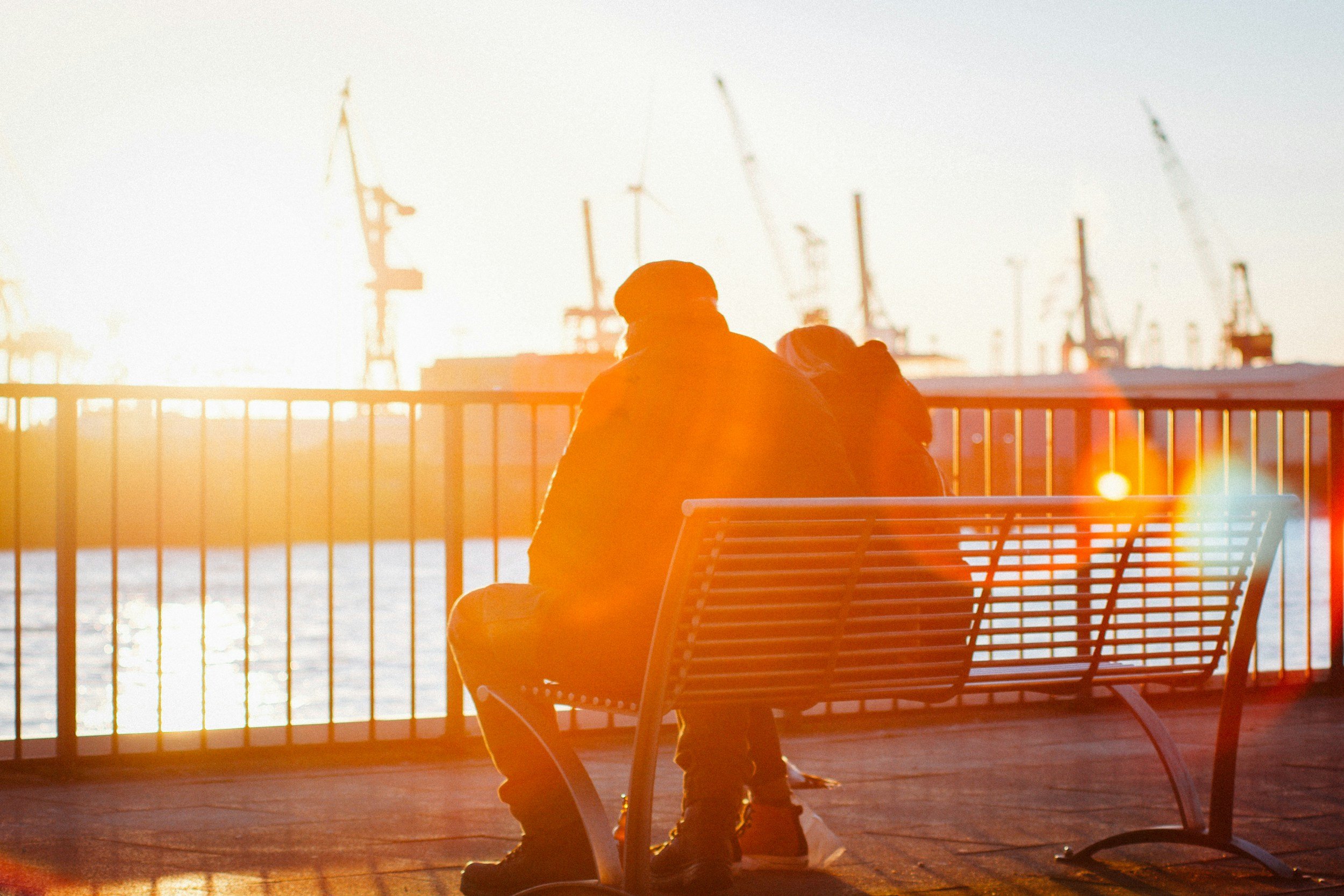 Person sitting on a bench near a waterfront during sunset or sunrise, with a fence and industrial cranes in the background.
