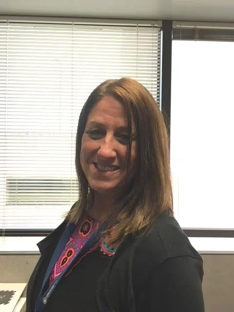 A professional photo of a woman with shoulder-length light brown hair, smiling in an office environment with large windows in the background.
