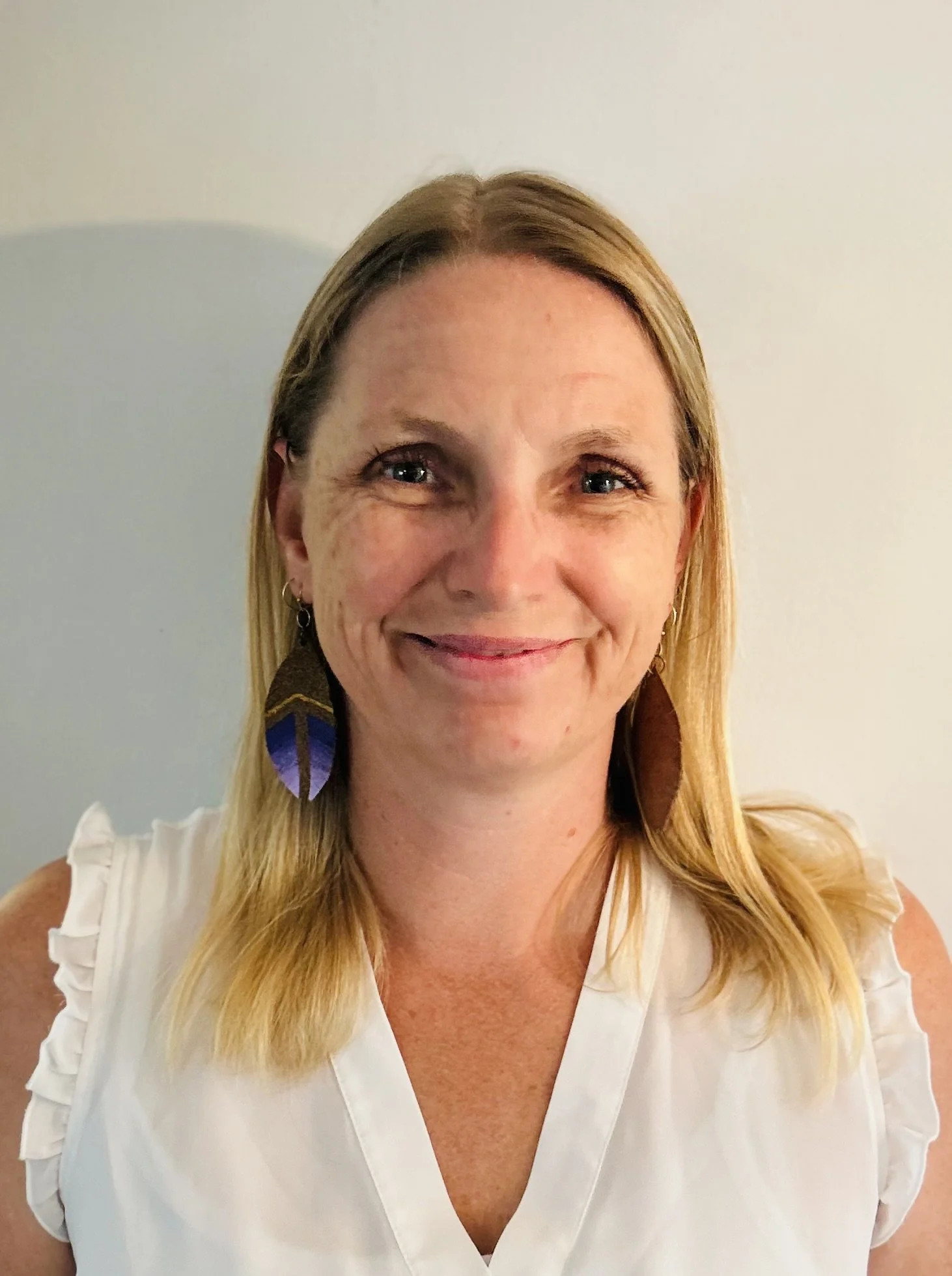 A portrait of a woman with straight blonde hair, wearing a sleeveless white top and dangly feather-shaped earrings, smiling gently against a light gray wall.
