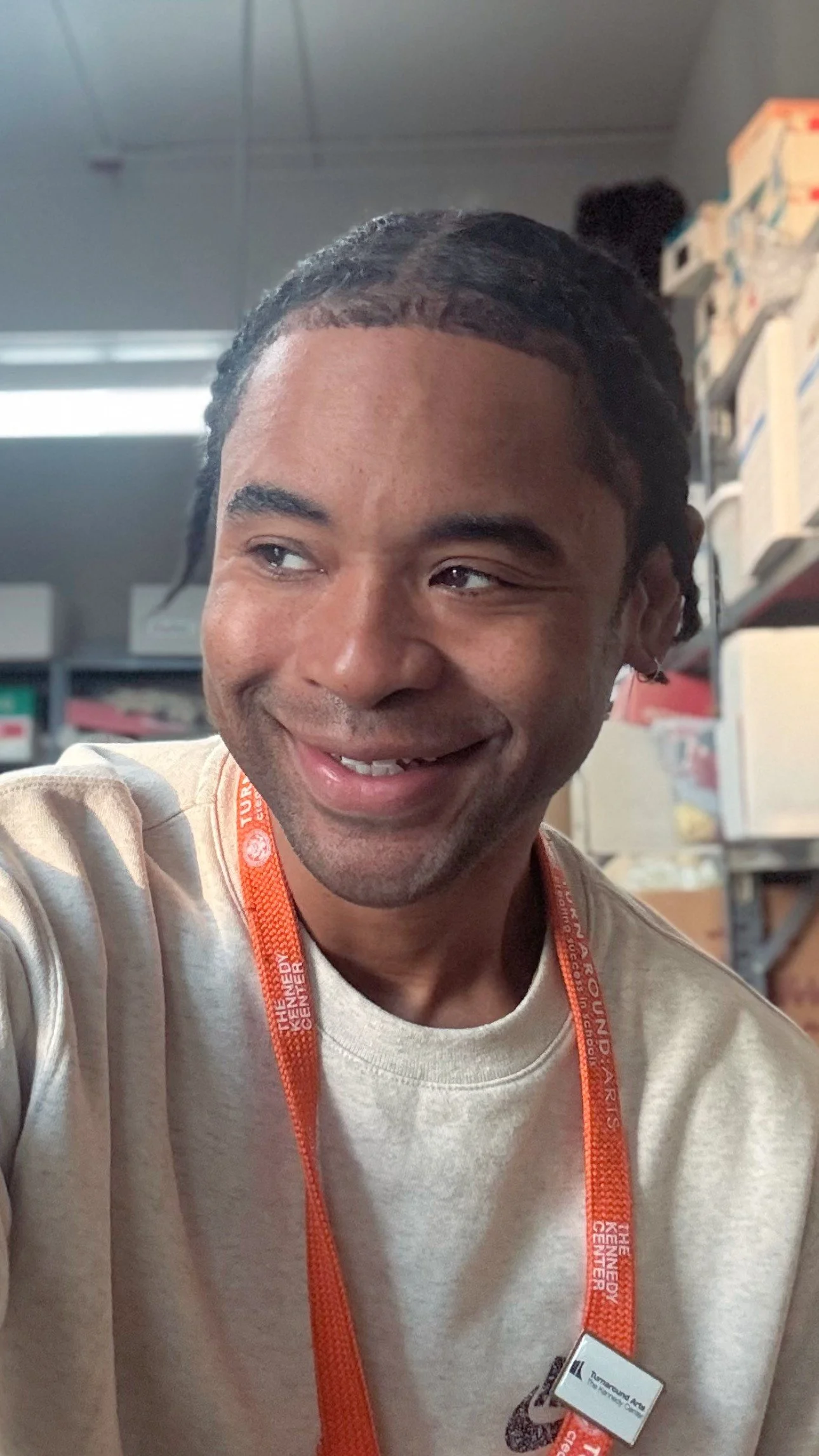A smiling individual with braided hair wearing a light-colored shirt and an orange lanyard in a room with shelves in the background.
