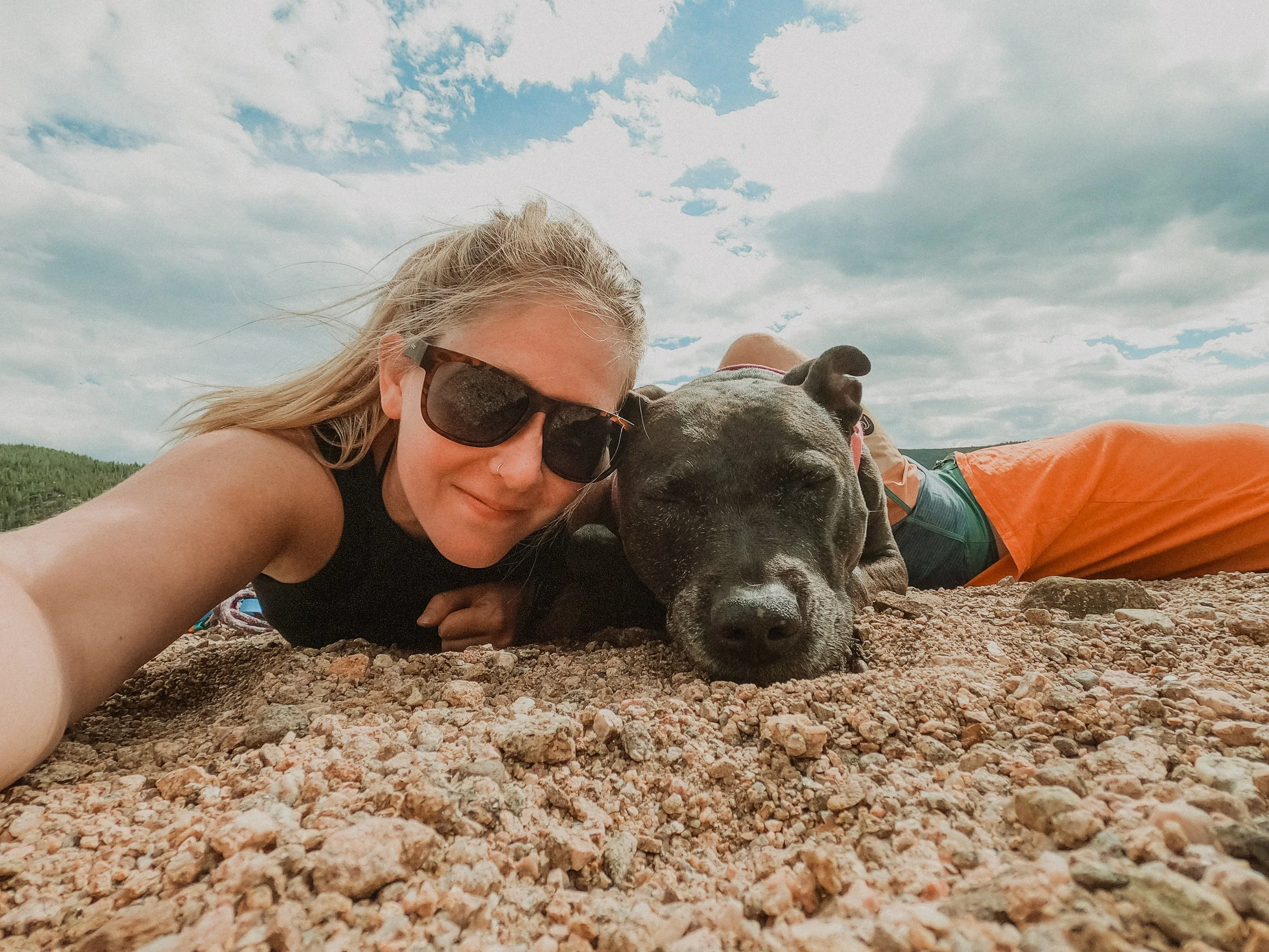 Woman with sunglasses lying on the ground next to a sleeping dog on a rocky surface under a cloudy sky.