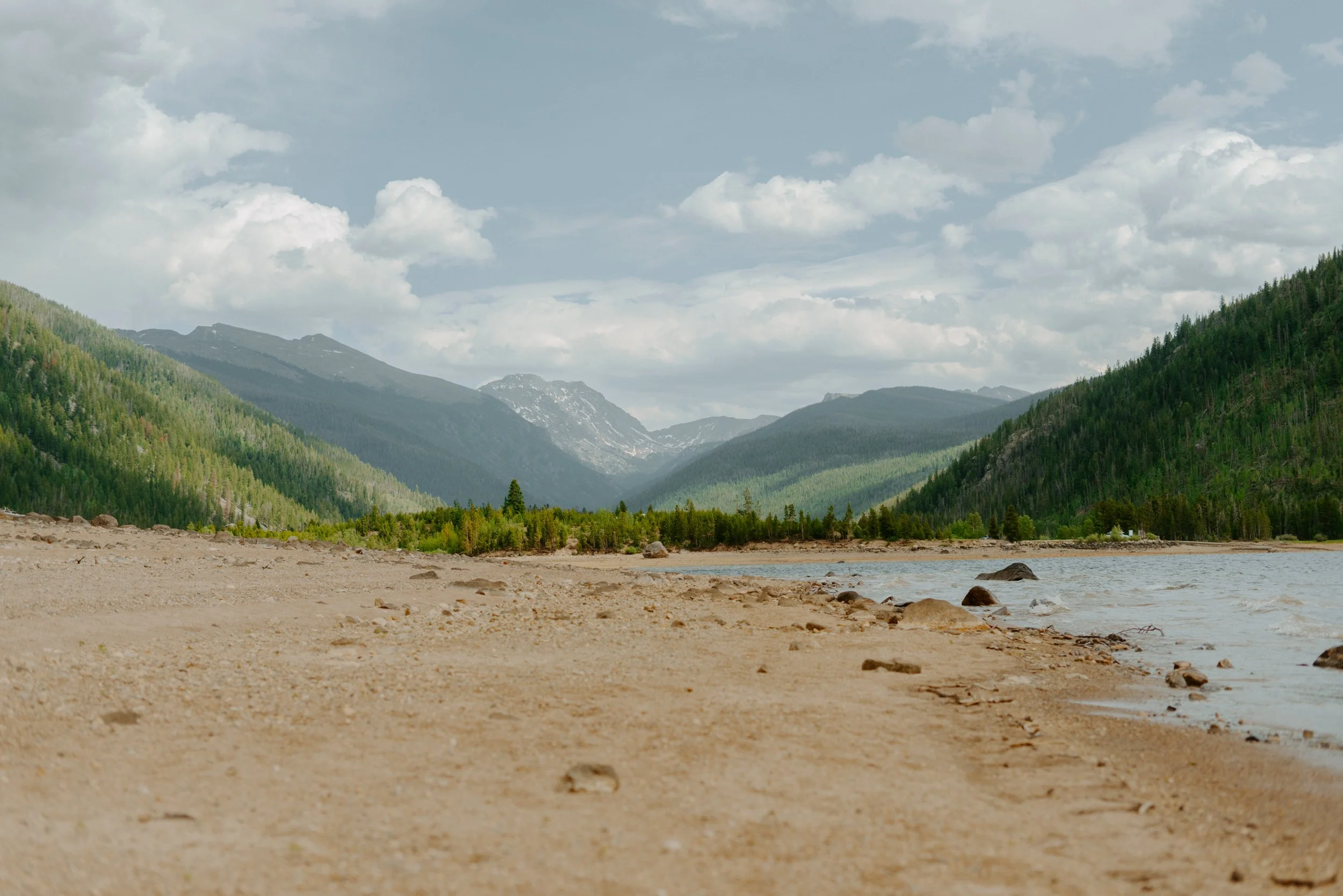 Mountain landscape with sandy shore, forested hills, and lake.