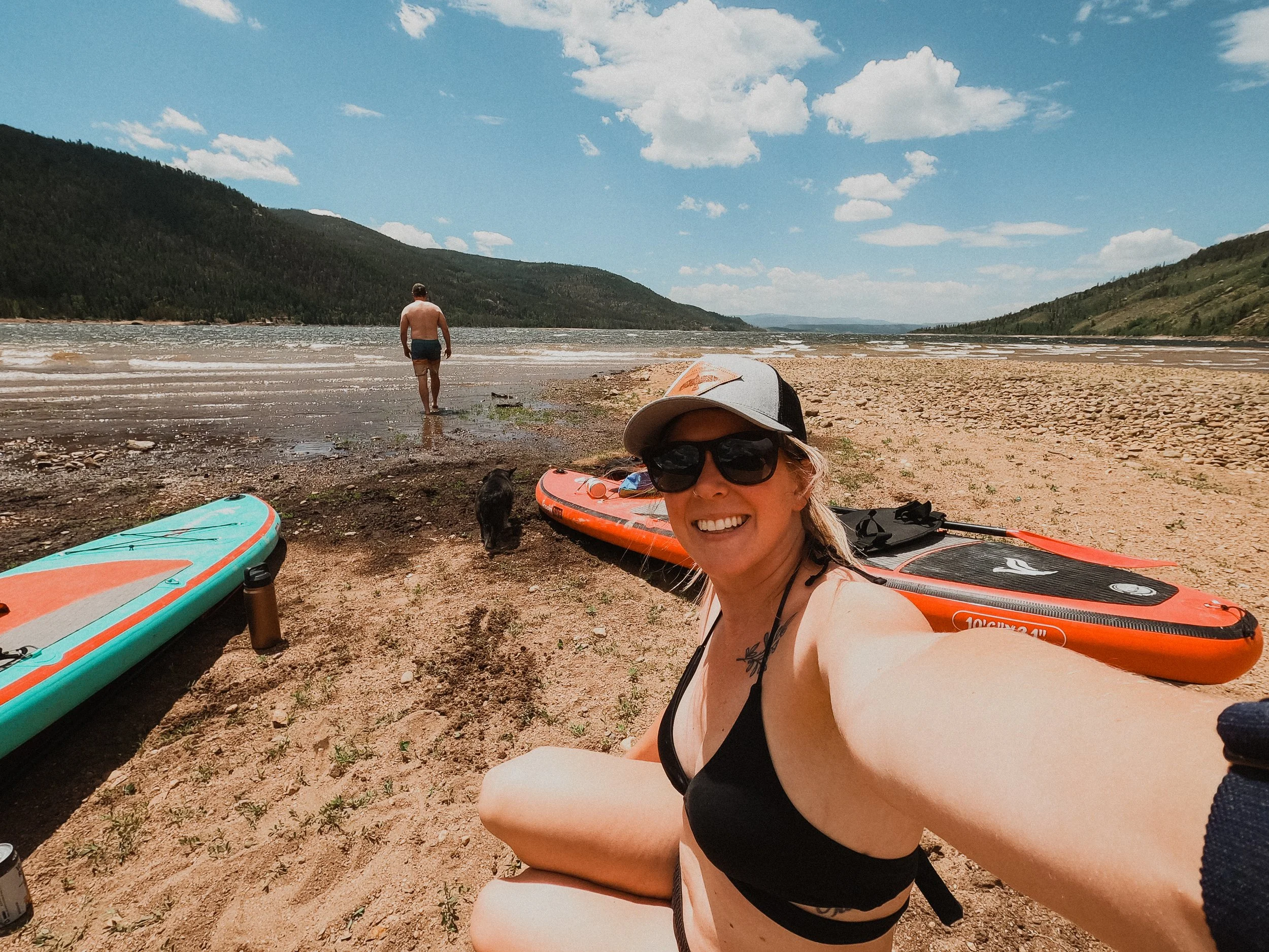 Woman taking a selfie on a lakeside beach with paddleboards, a man walking towards the water, mountainous scenery, and a dog nearby.