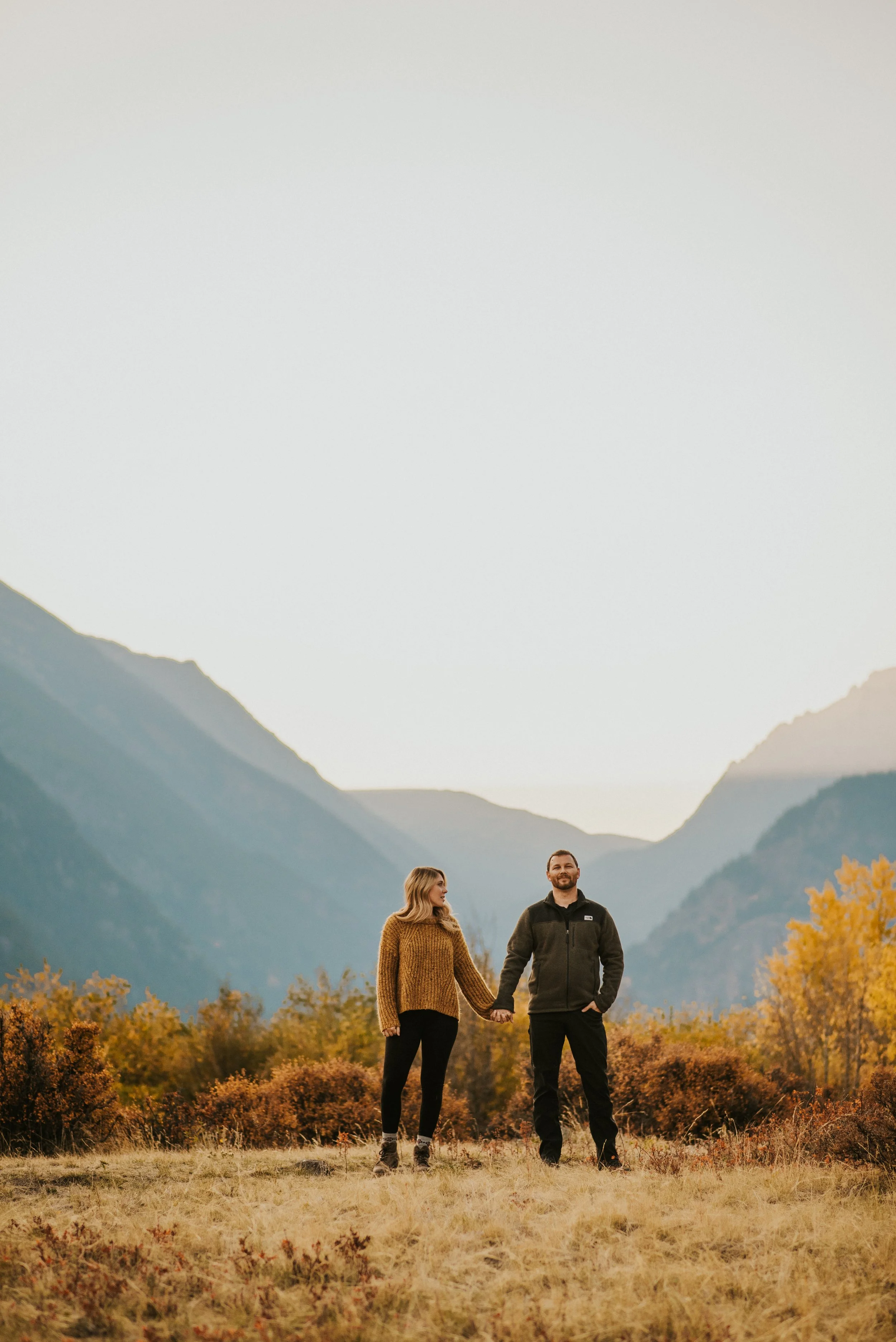 Couple holding hands in a grassy field with mountains in the background, surrounded by autumn foliage.