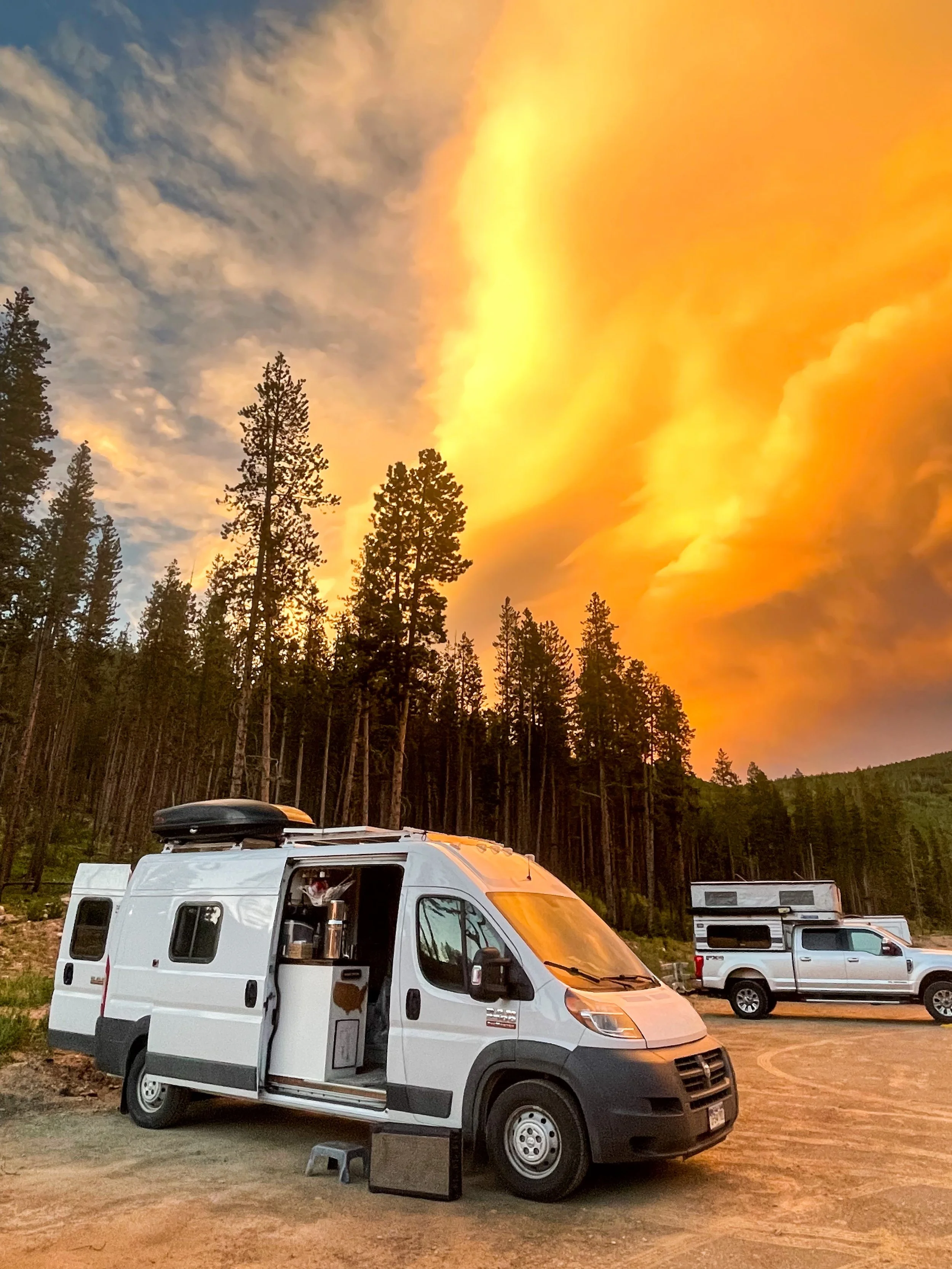 White camper van parked in a wooded area at sunset with orange clouds in the sky.