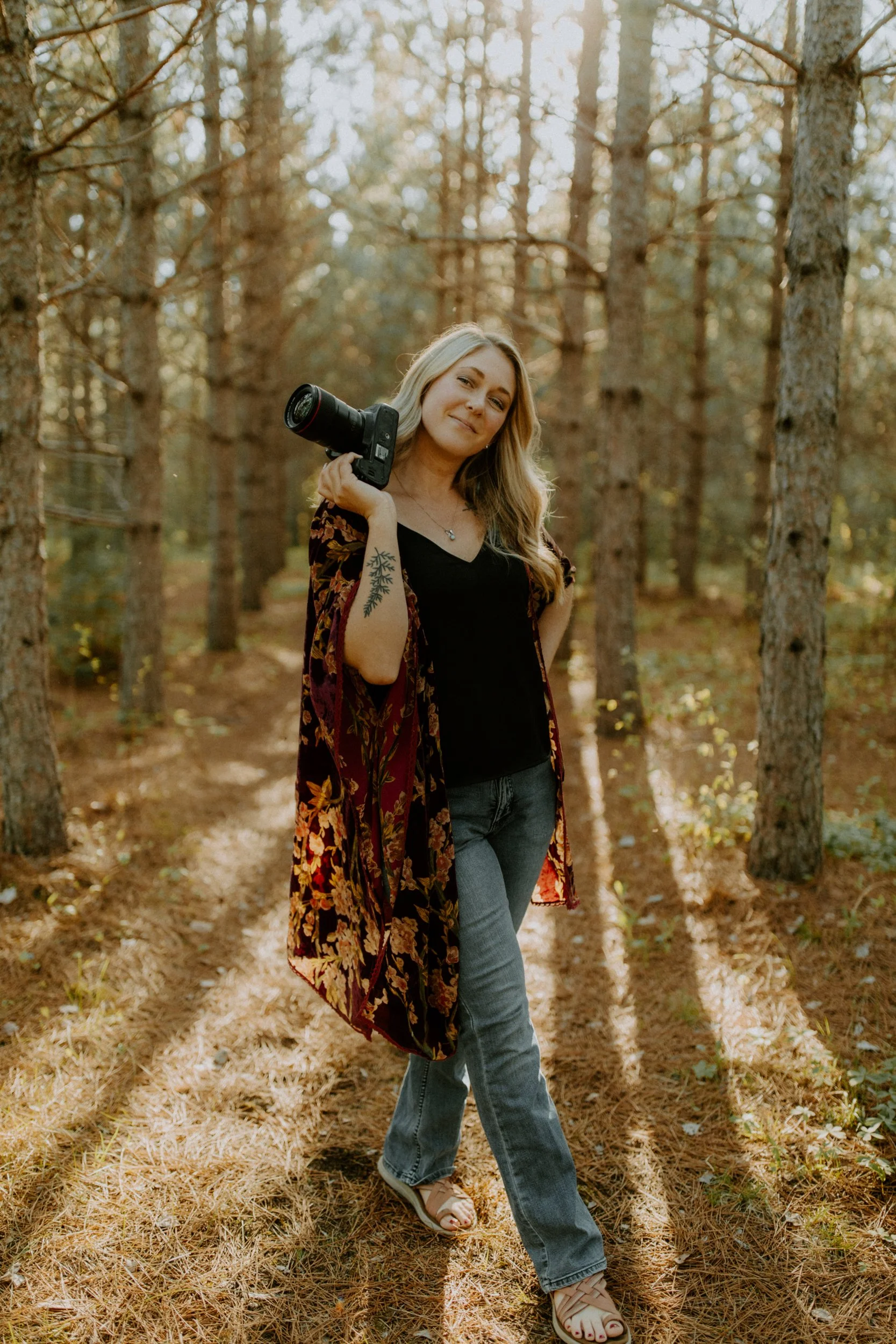 Woman holding a camera over her shoulder, standing in a sunlit forest, wearing a floral shawl, black top, and jeans.