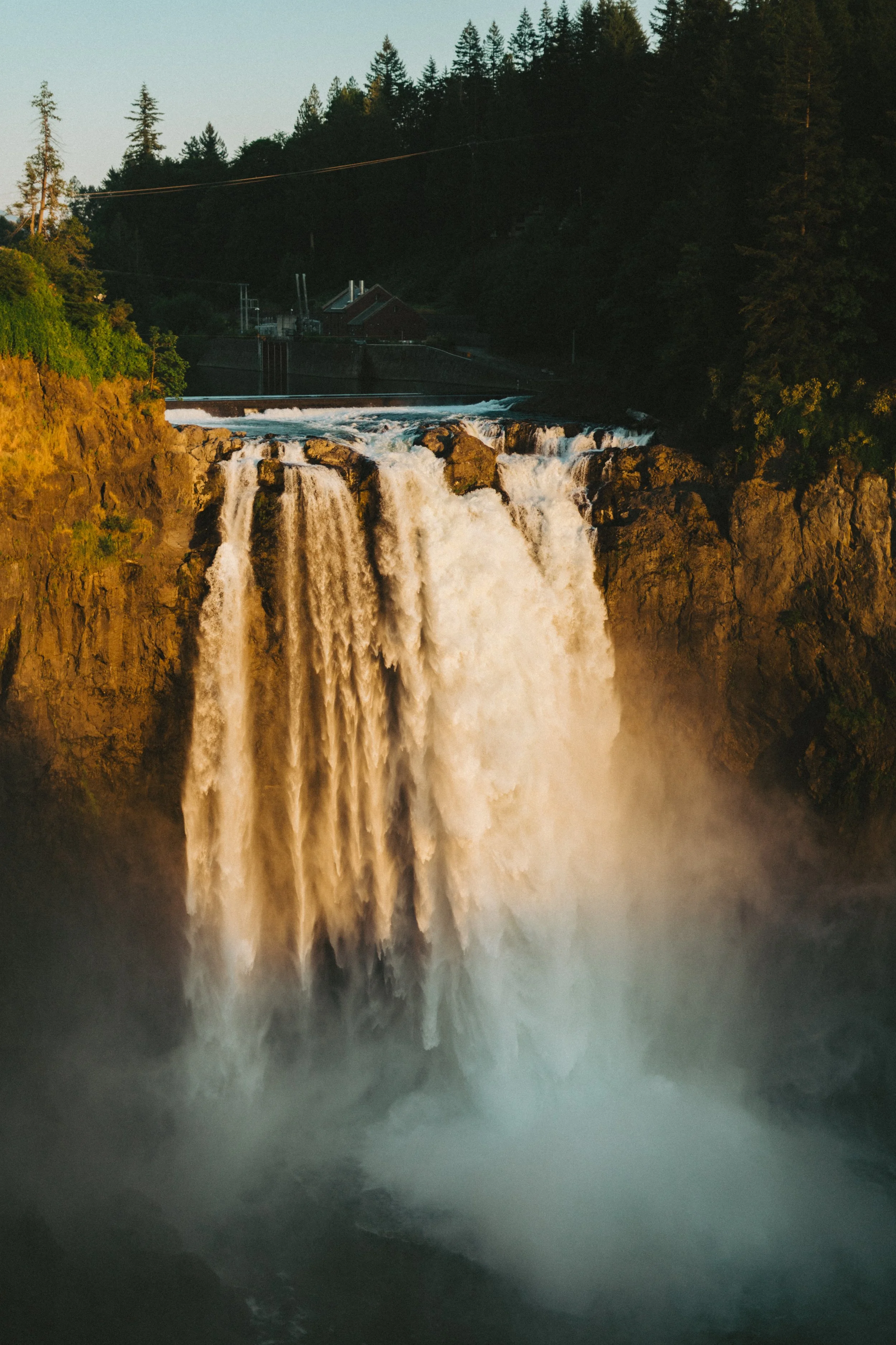 Scenic waterfall cascading over rocks with lush green forest and a small building in the background at sunset.
