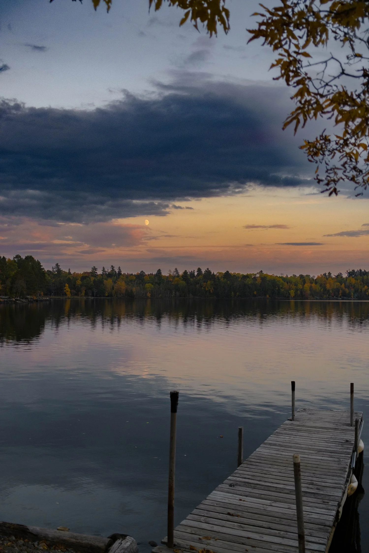 A wooden dock extends over a calm lake under a partly cloudy sunset sky, with trees lining the opposite shore and the moon visible.