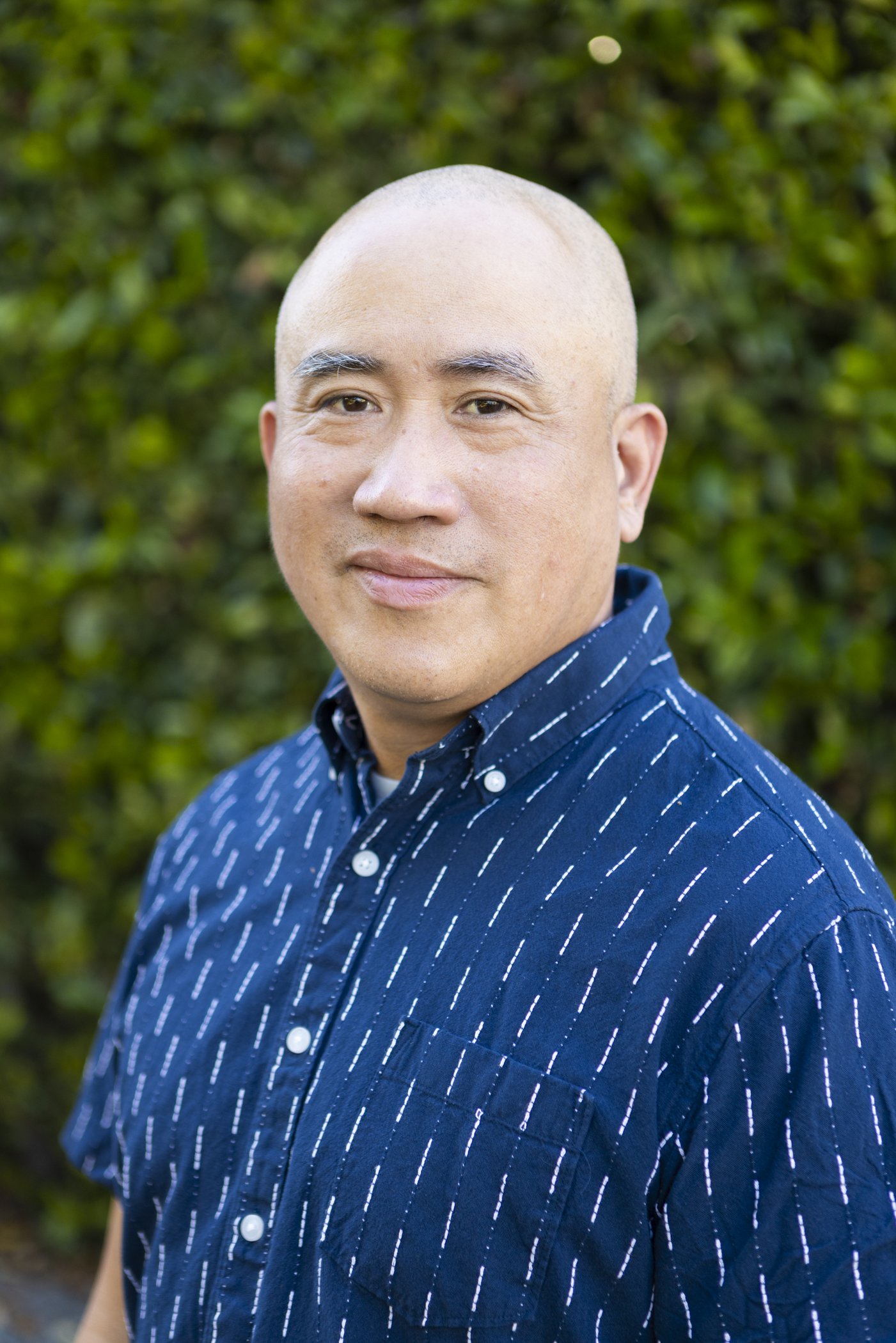 Close-up portrait of a middle-aged Asian man with a shaved head, wearing a dark blue button-up shirt with white stripes, standing outdoors with green foliage in the background.