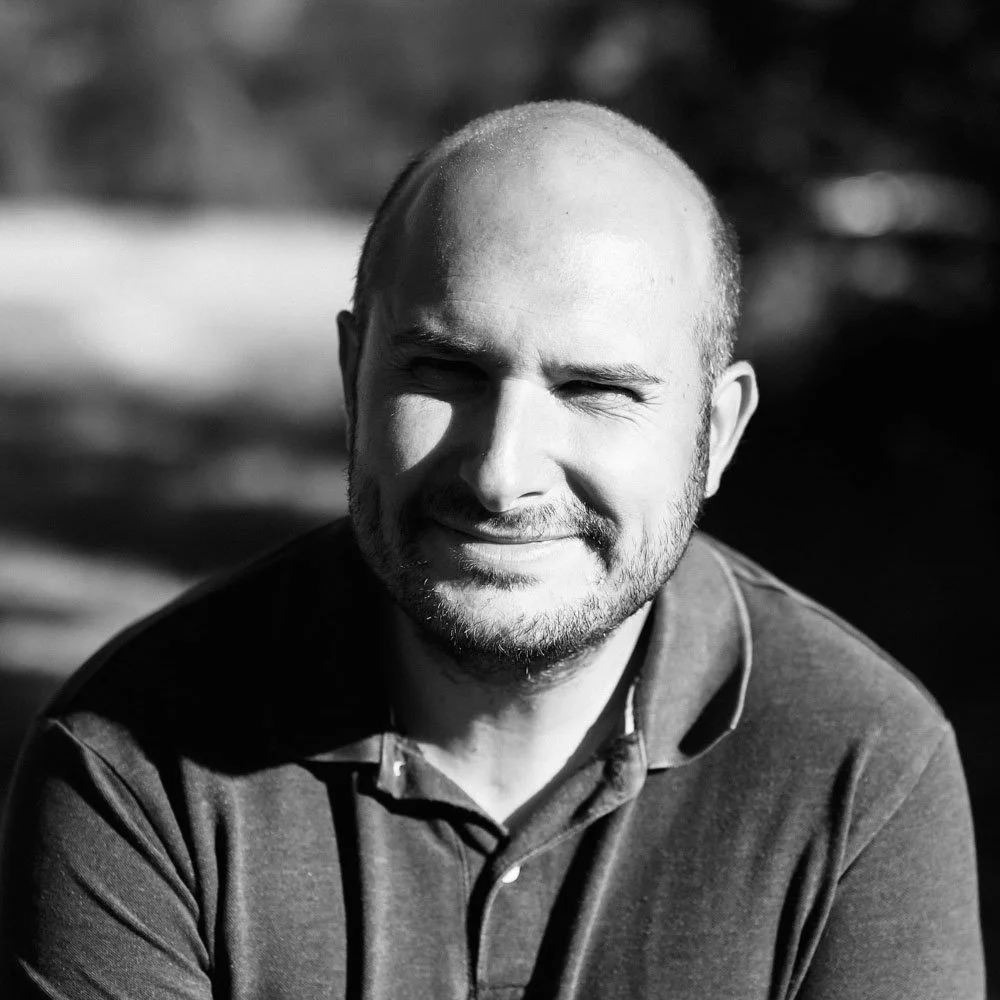 Portrait en noir et blanc d'un homme souriant avec un visage chauve et une barbe, portant un pull, en plein air avec un fond flou.