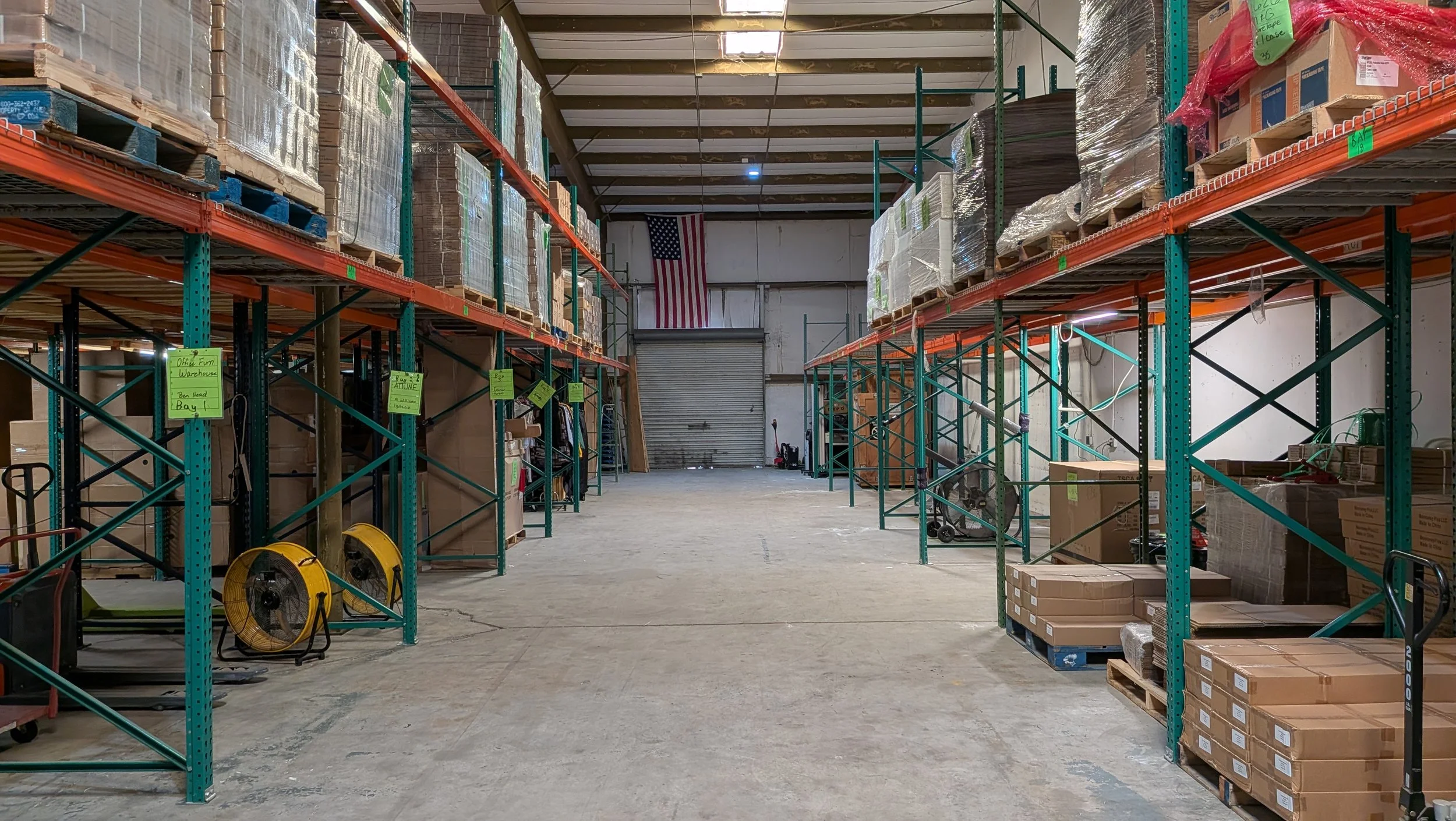 Empty warehouse with tall shelves stocked with boxes and pallets, American flag hanging on back wall.