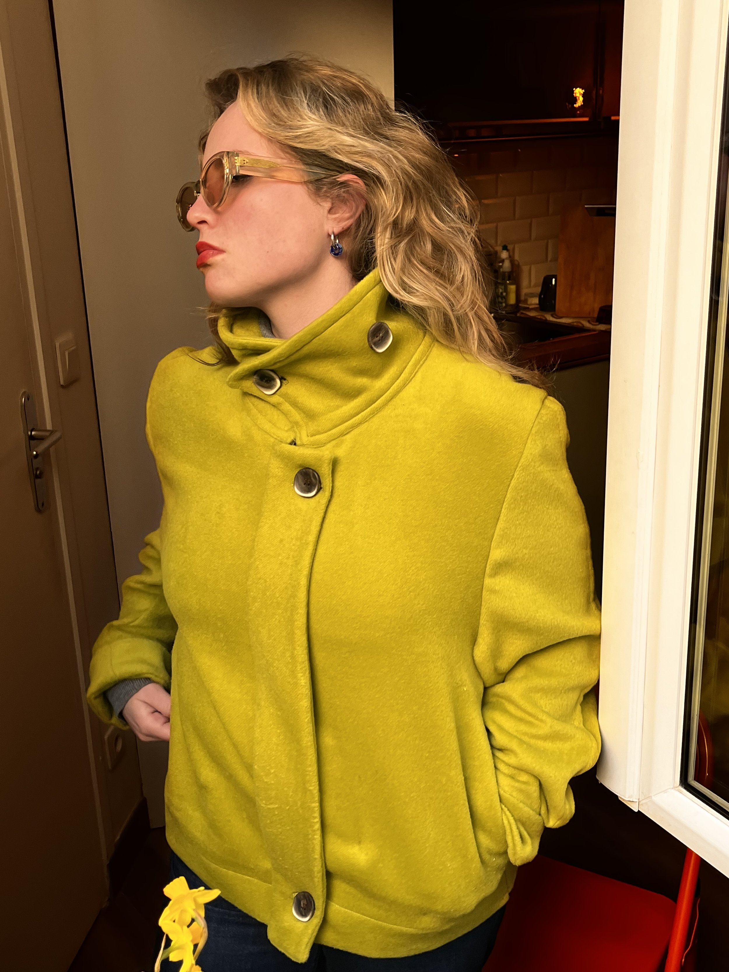 Photo d’une jeune femme blonde à la fenêtre d’un appartement parisien. Elle porte une veste verte couleur vert anis et cousue à la main