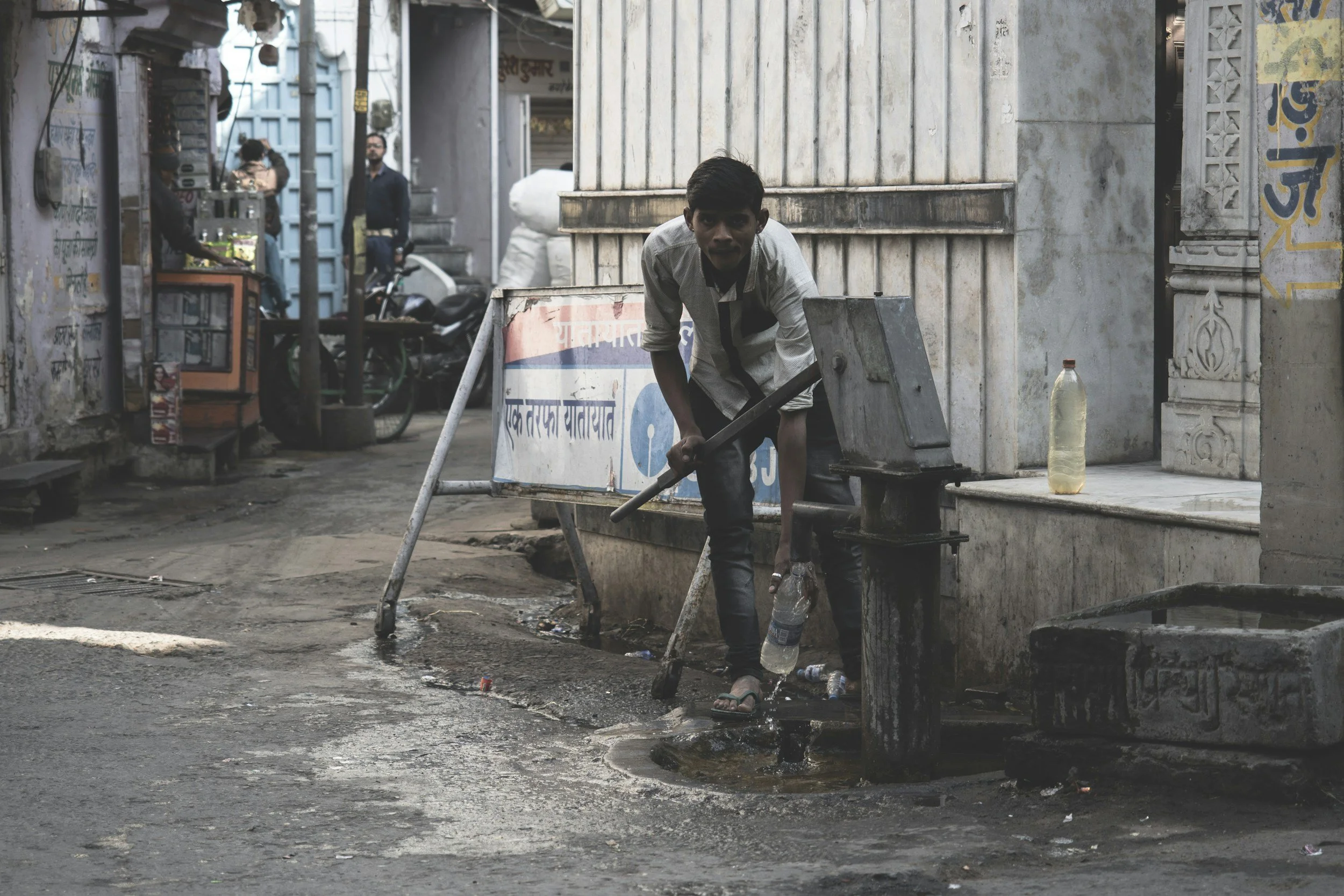 A young man in casual clothes pumping water from a street water source in an urban area.
