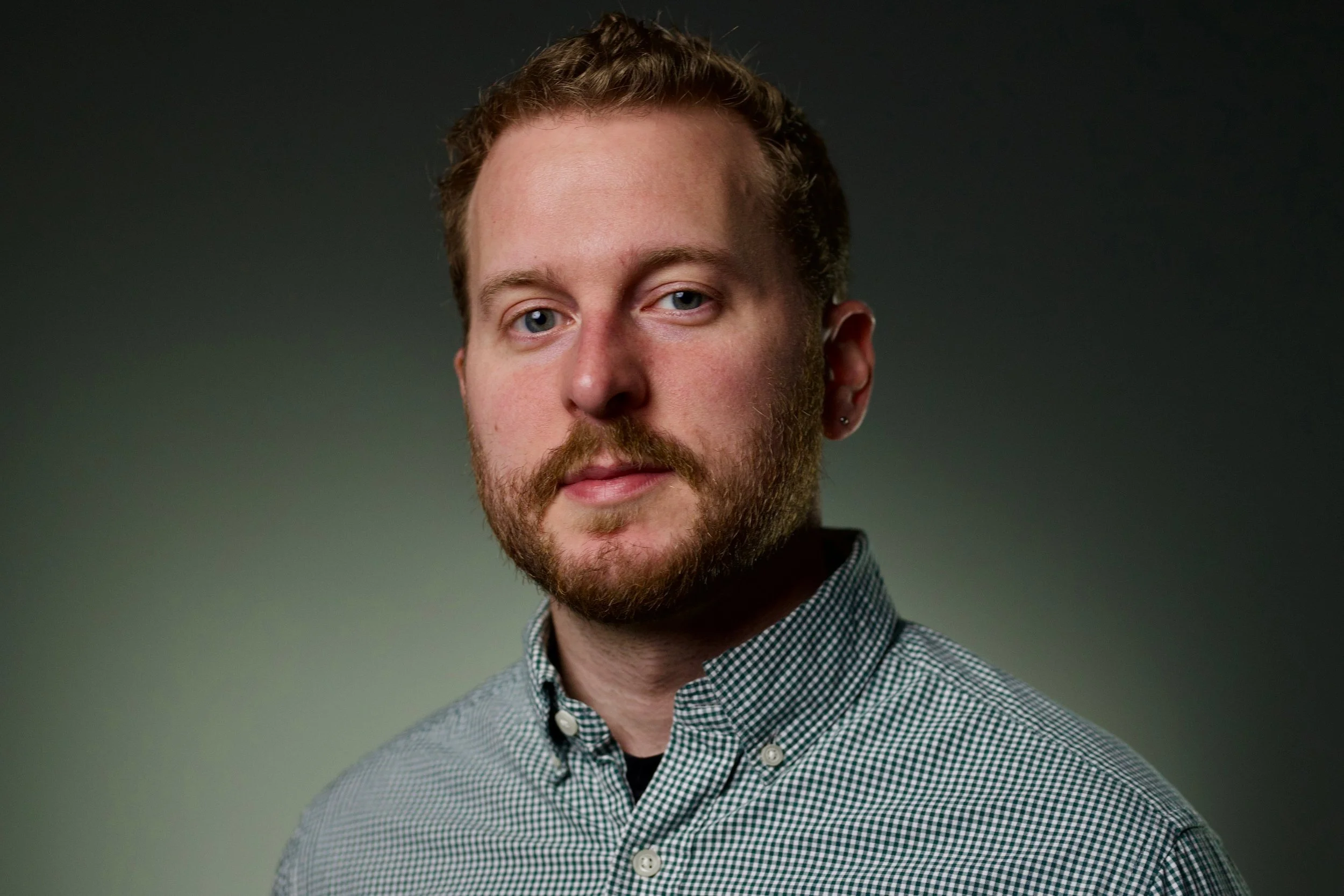 Headshot of a man with light skin, light brown hair, beard, wearing a black and white checkered collared shirt, against a dark gradient background.