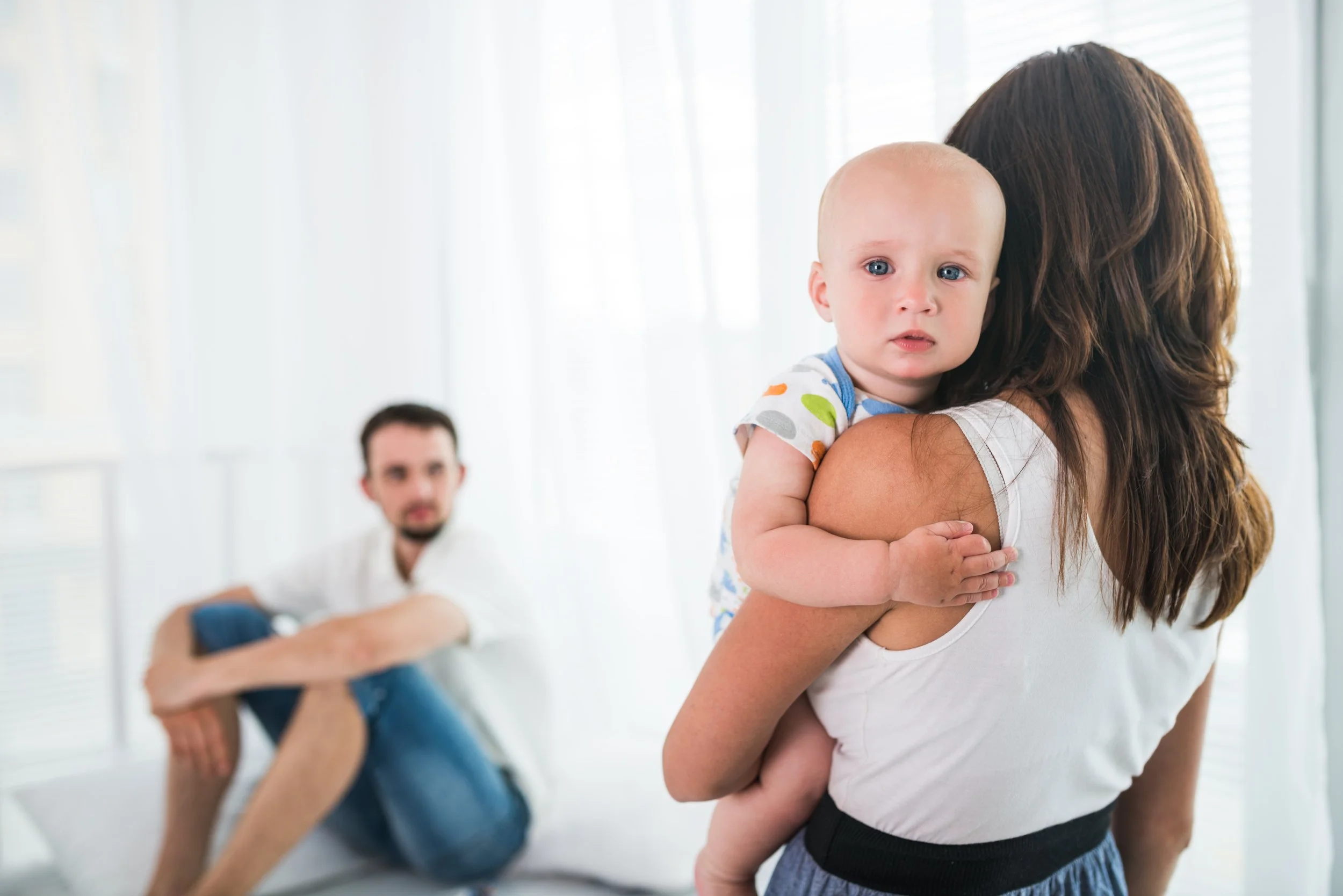A mother holding a baby while a father sits apart in the background, both exhausted and disconnected after having a child