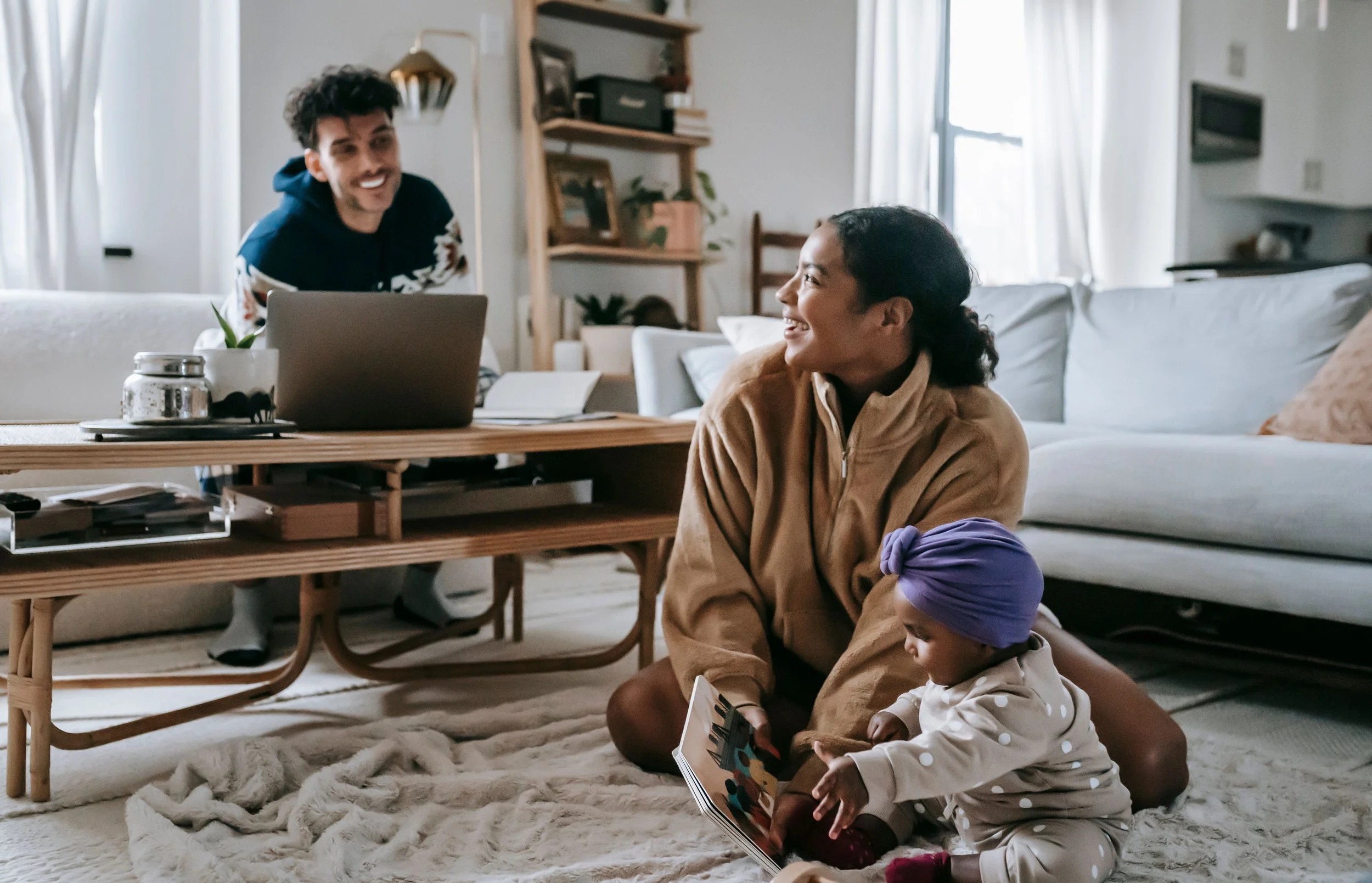 Parents smiling at each other while one plays with the baby and the other works nearby, reconnecting in an ordinary moment at home