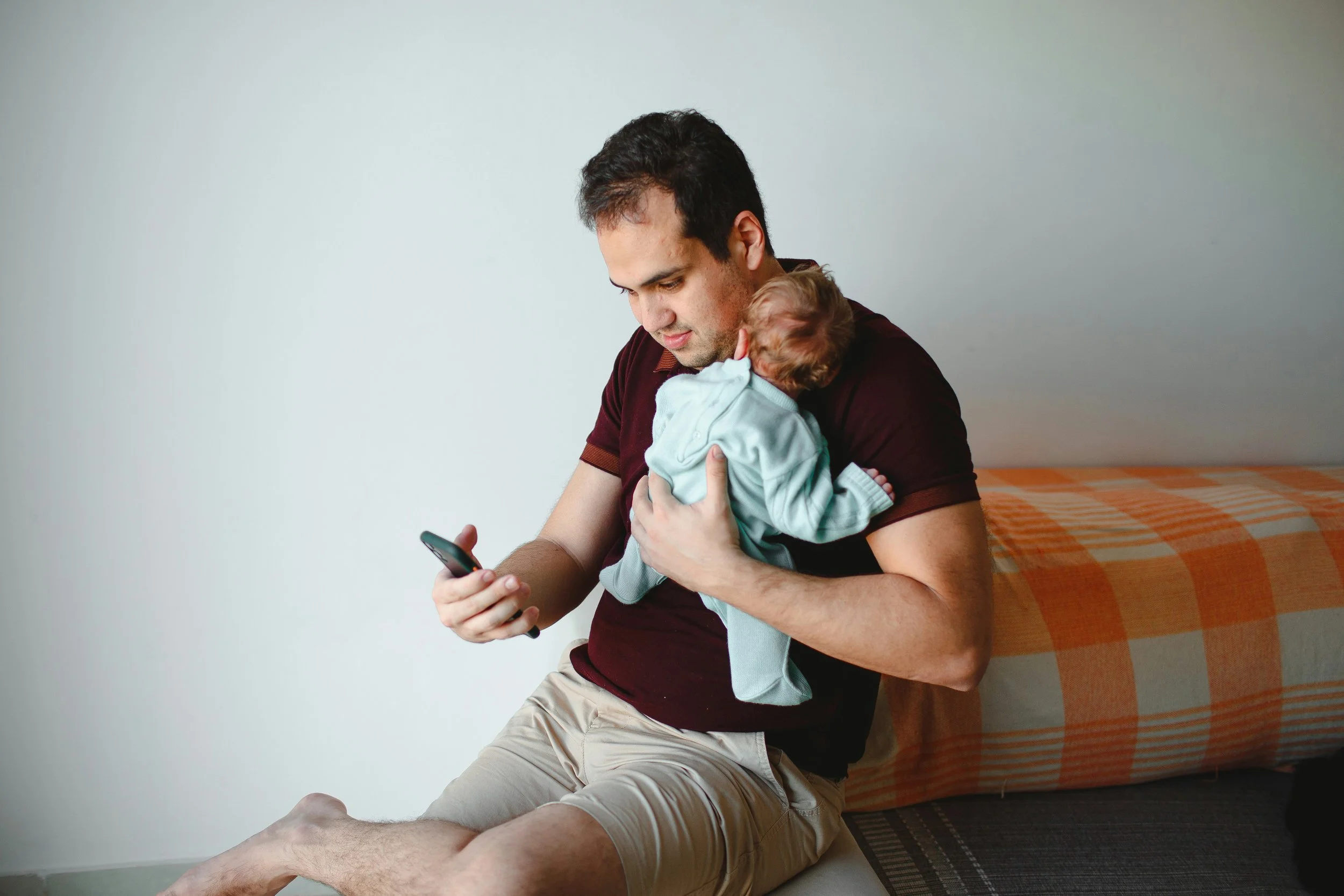 A father holding his baby while looking at his phone showing the pattern of staying busy and functional that can mask paternal postpartum depression.
