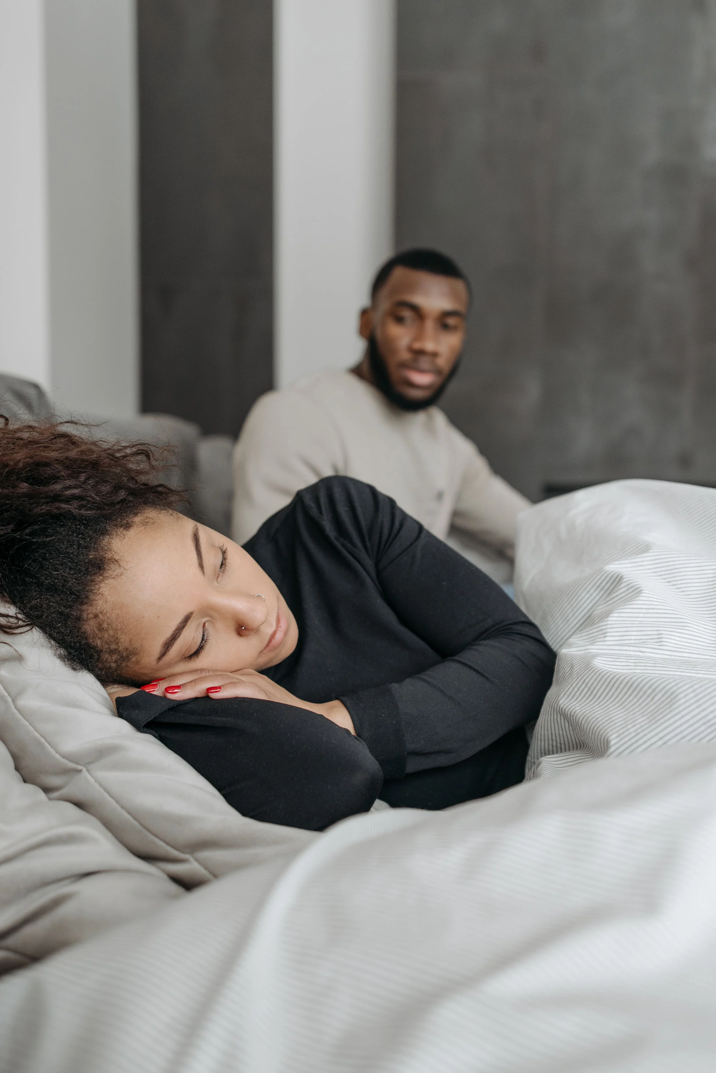 Couple lying apart in bed, woman turned away and man sitting at a distance, reflecting the physical and emotional disconnection around intimacy after having a baby
