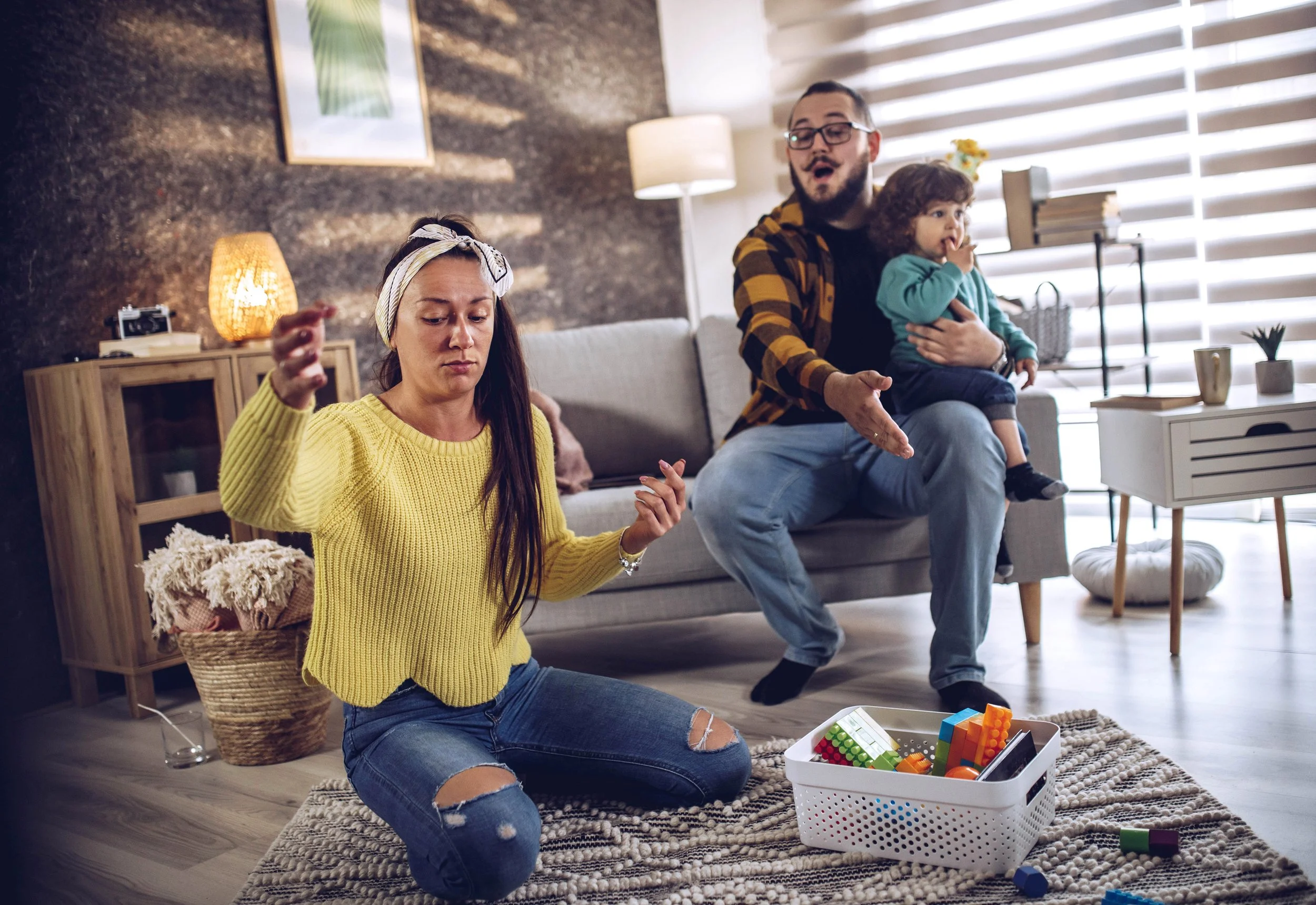 A woman sitting on the floor looking frustrated while her partner holds their baby on the couch behind her, showing the disconnection that criticism creates between new parents.