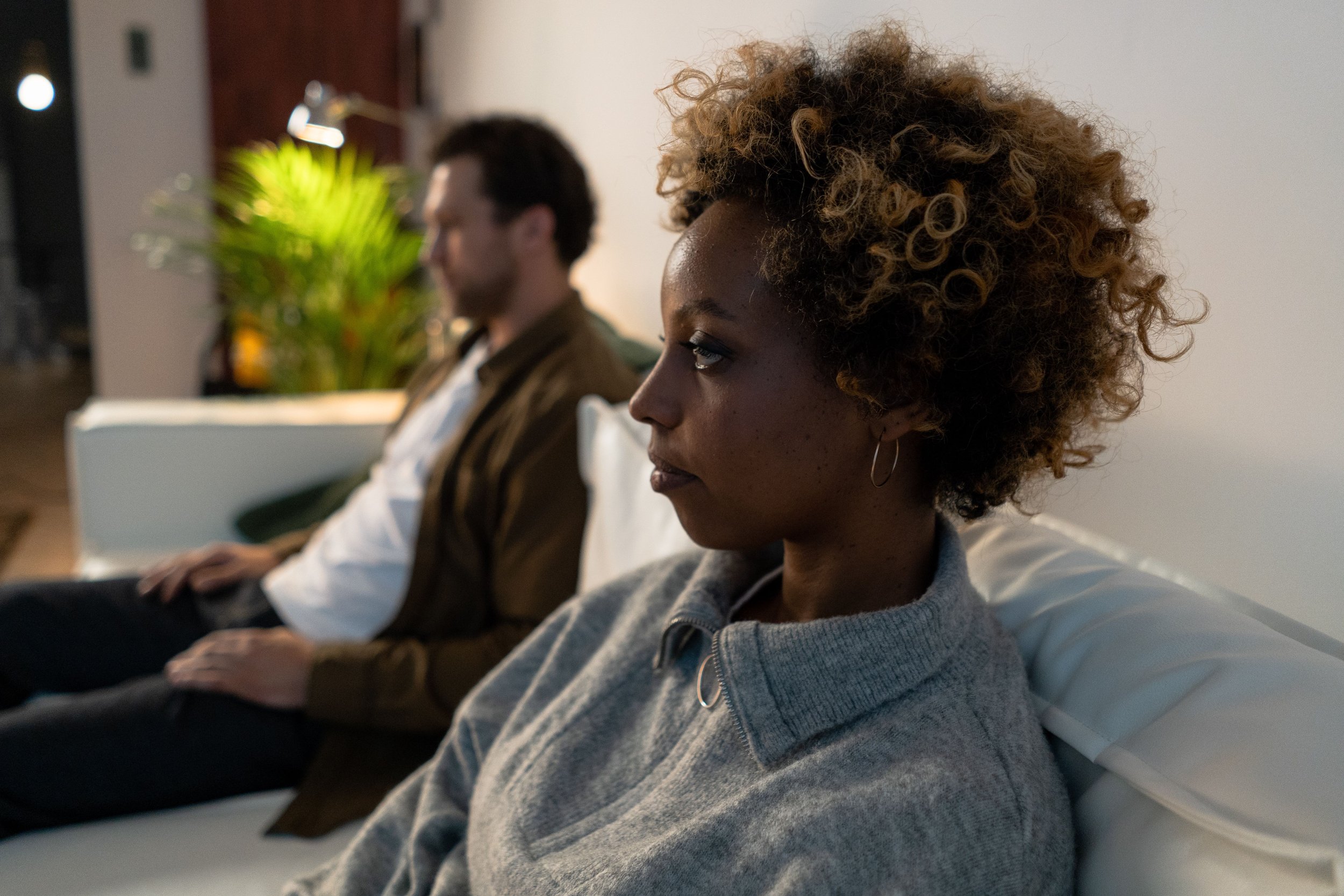 Couple sitting on the same couch looking in opposite directions, capturing the quiet distance and sense of growing apart that many couples experience after having a baby