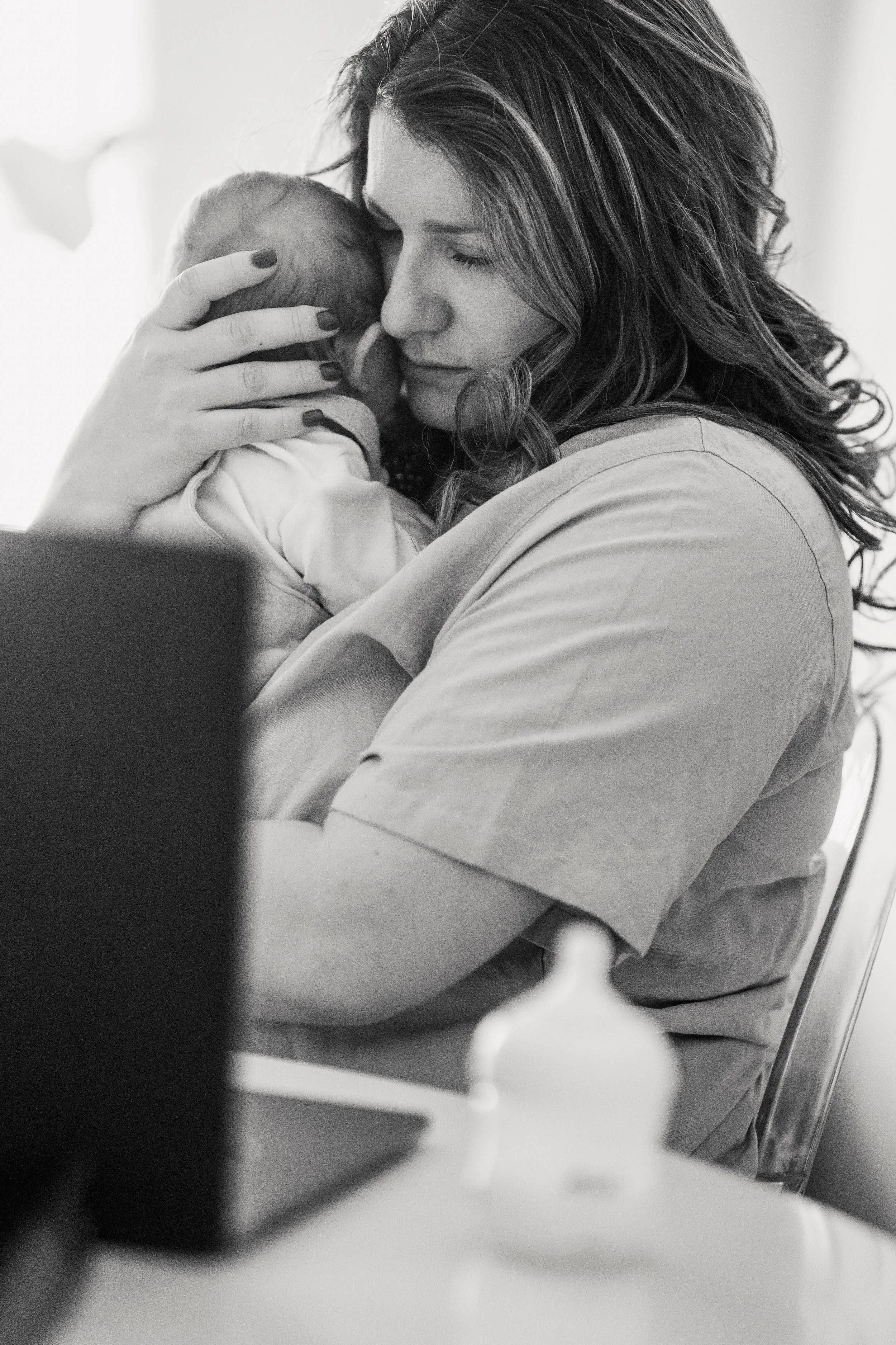 Exhausted new mother holding her baby while trying to work, showing the depletion and invisible load of early postpartum