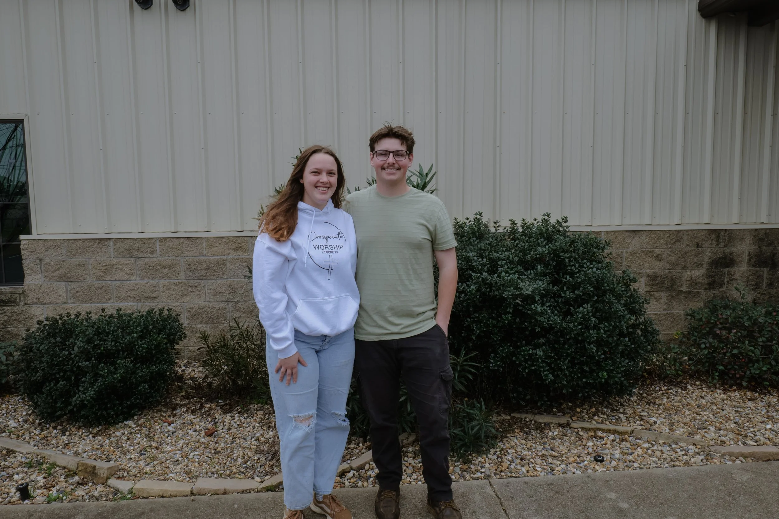 Two young adults, a woman wearing a white hoodie and ripped jeans, and a man in a striped t-shirt and dark pants, standing outside in front of a beige metal building with bushes and rocks around them. They are smiling at the camera.